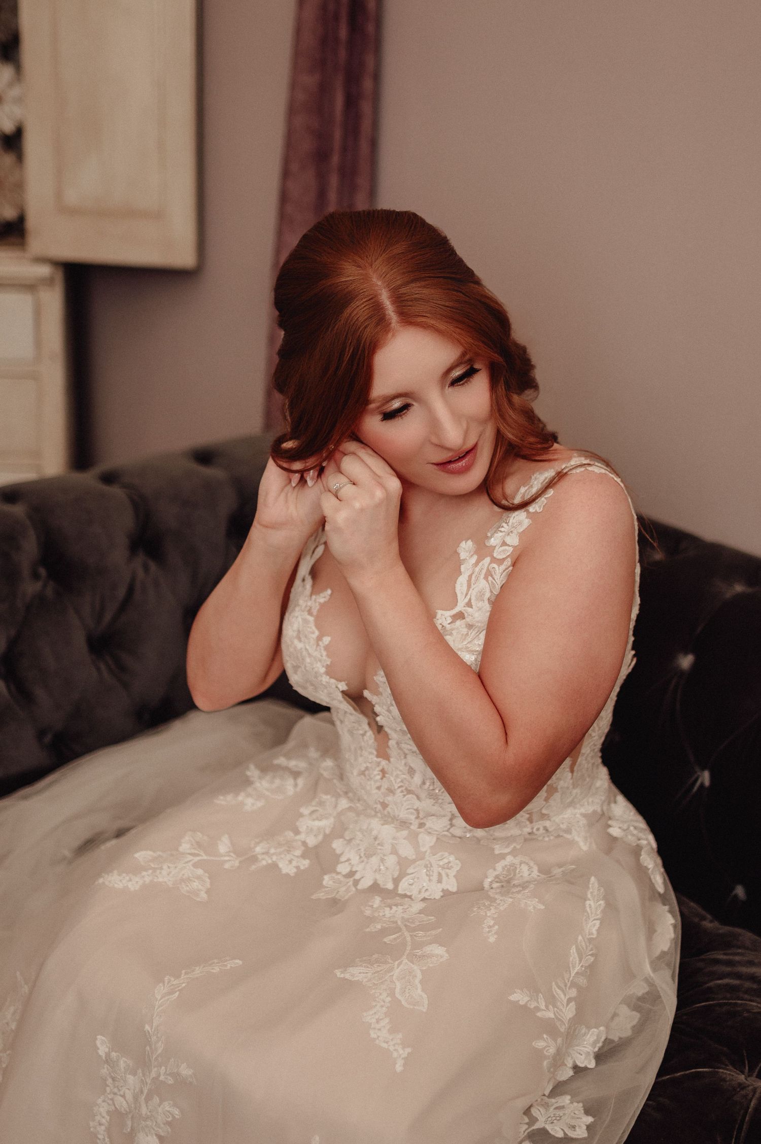 A bride in a white lace wedding dress with floral details poses elegantly on a dark couch.
