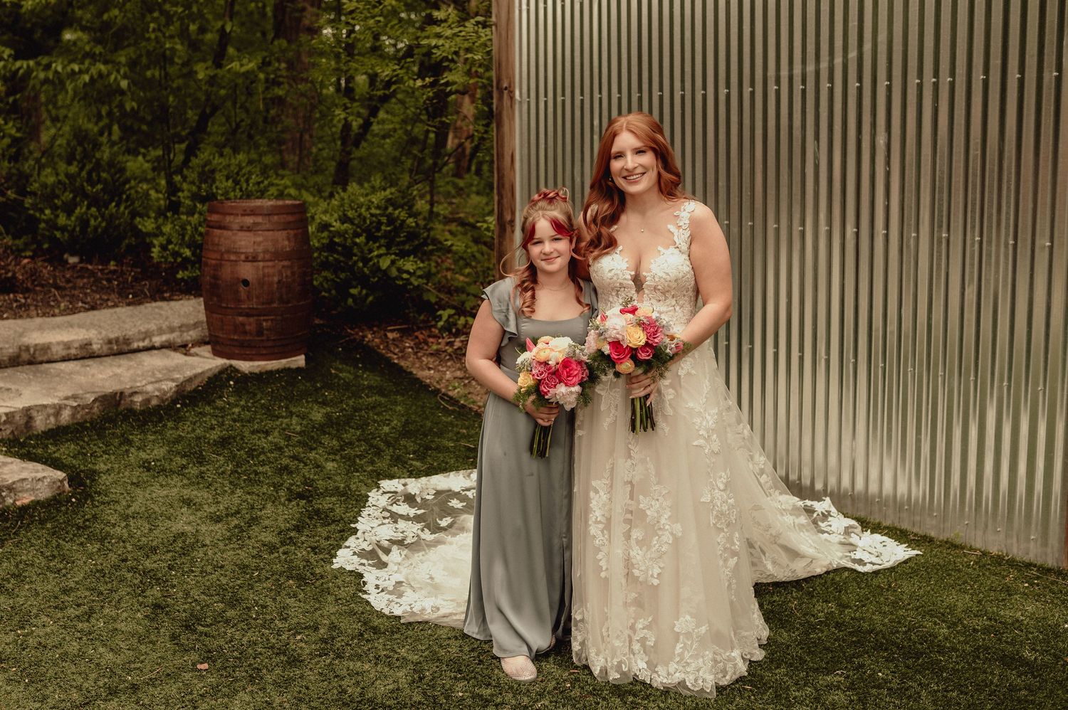 A wedding party poses in front of a rustic corrugated metal wall with lush greenery in the background.