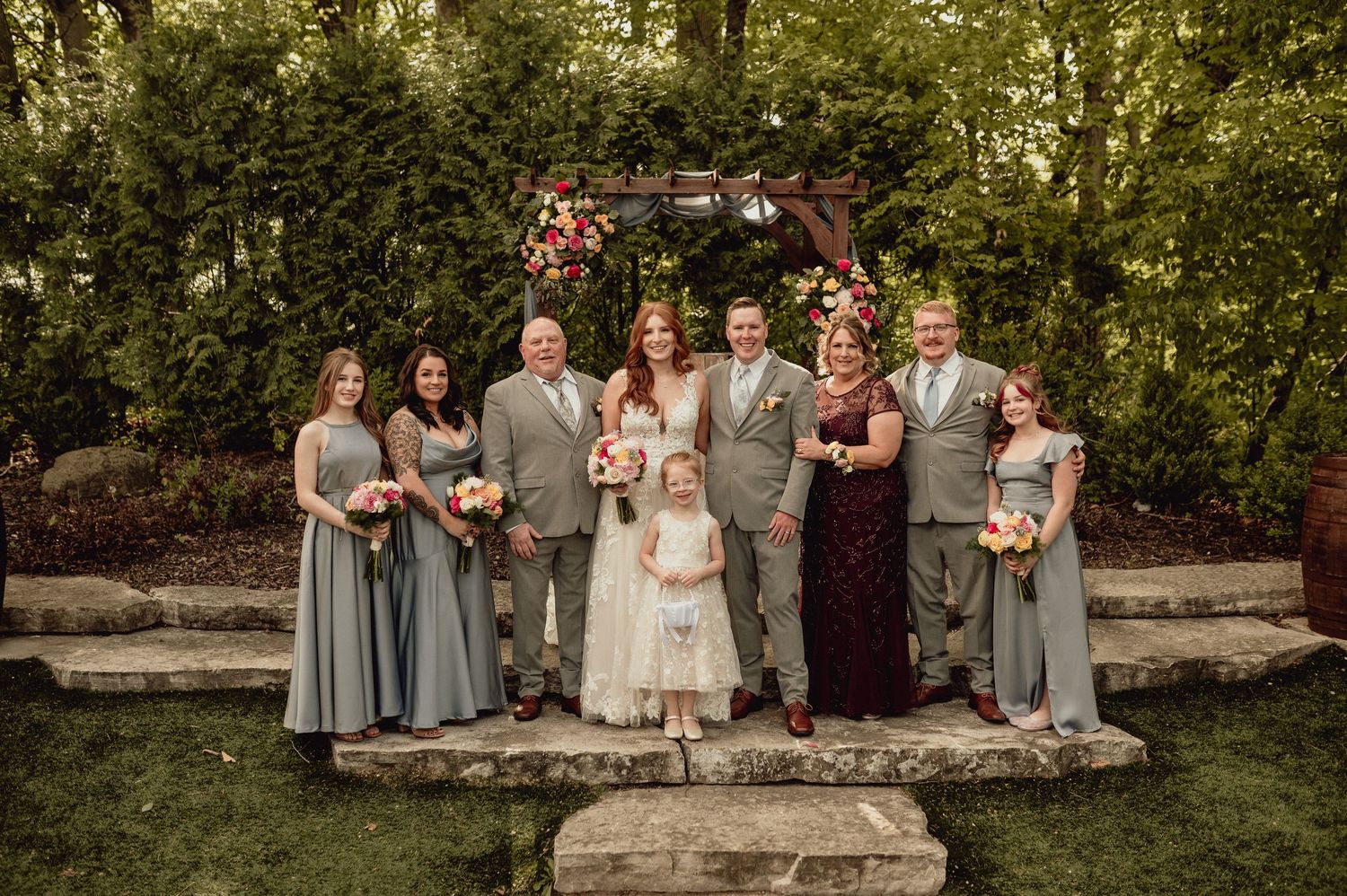 A wedding party poses together on stone steps in a lush garden setting during golden hour.