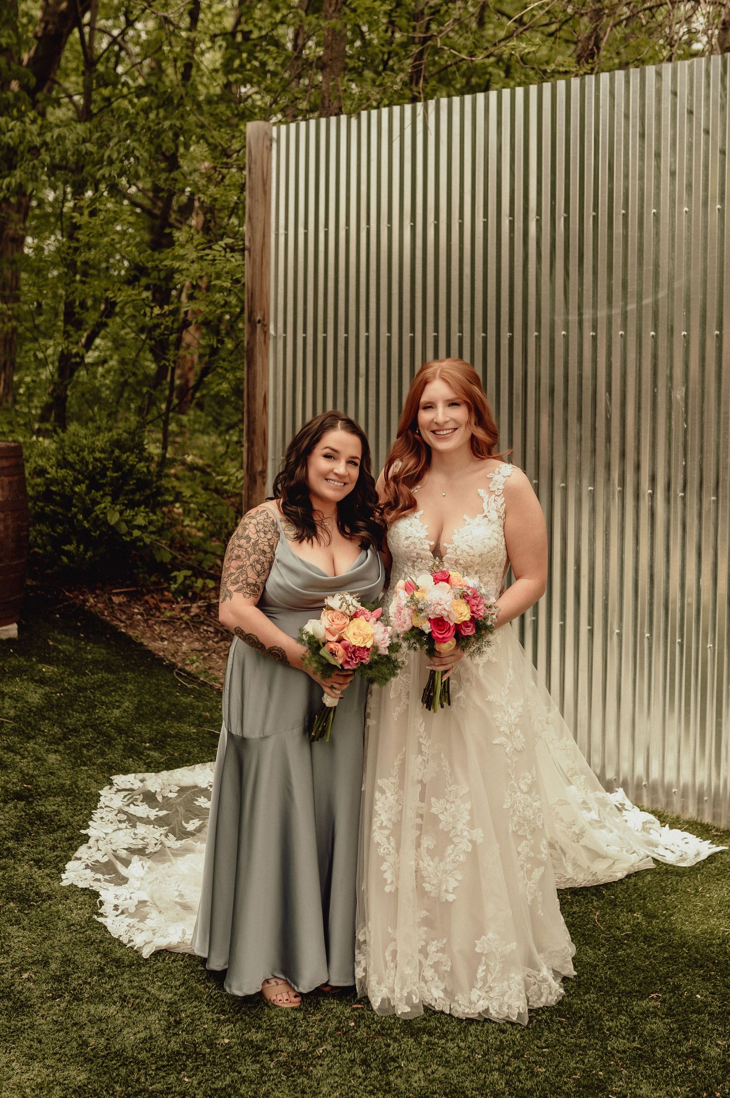 Two wedding attendees pose together outdoors against a white fence, one wearing a gray dress and one in a floral gown.