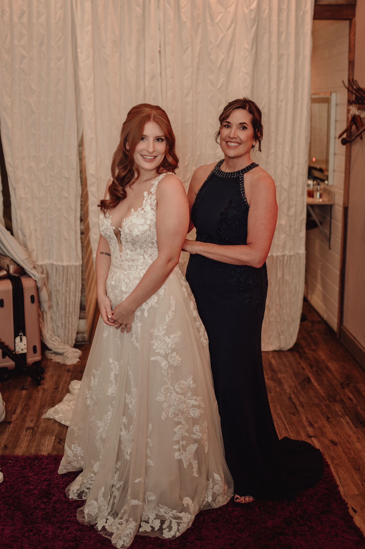 Two people pose together in formal attire at an indoor venue with wooden floors and white curtains in the background.
