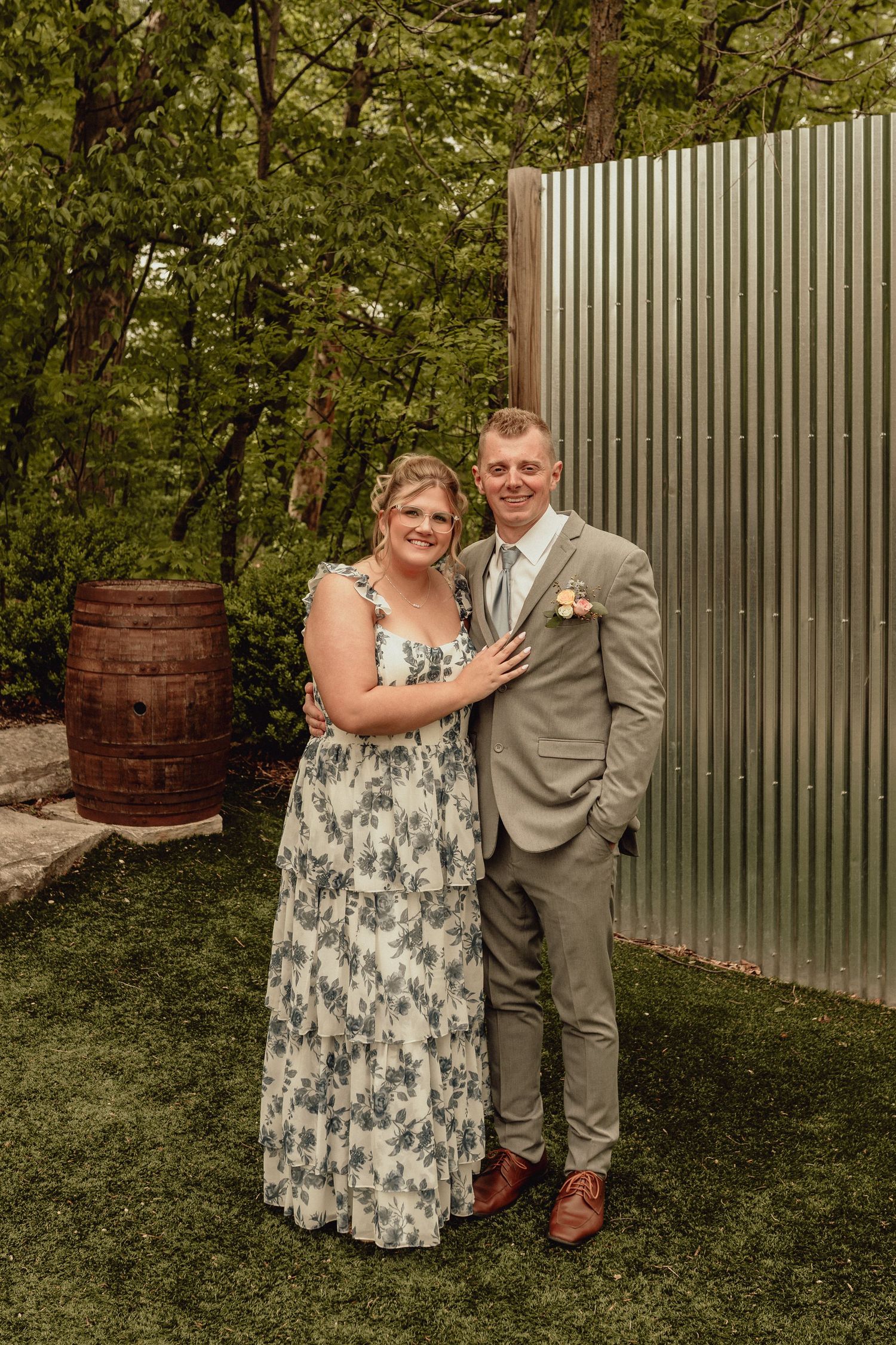 A couple poses together outdoors near a wooden barrel and metal wall in a garden setting.