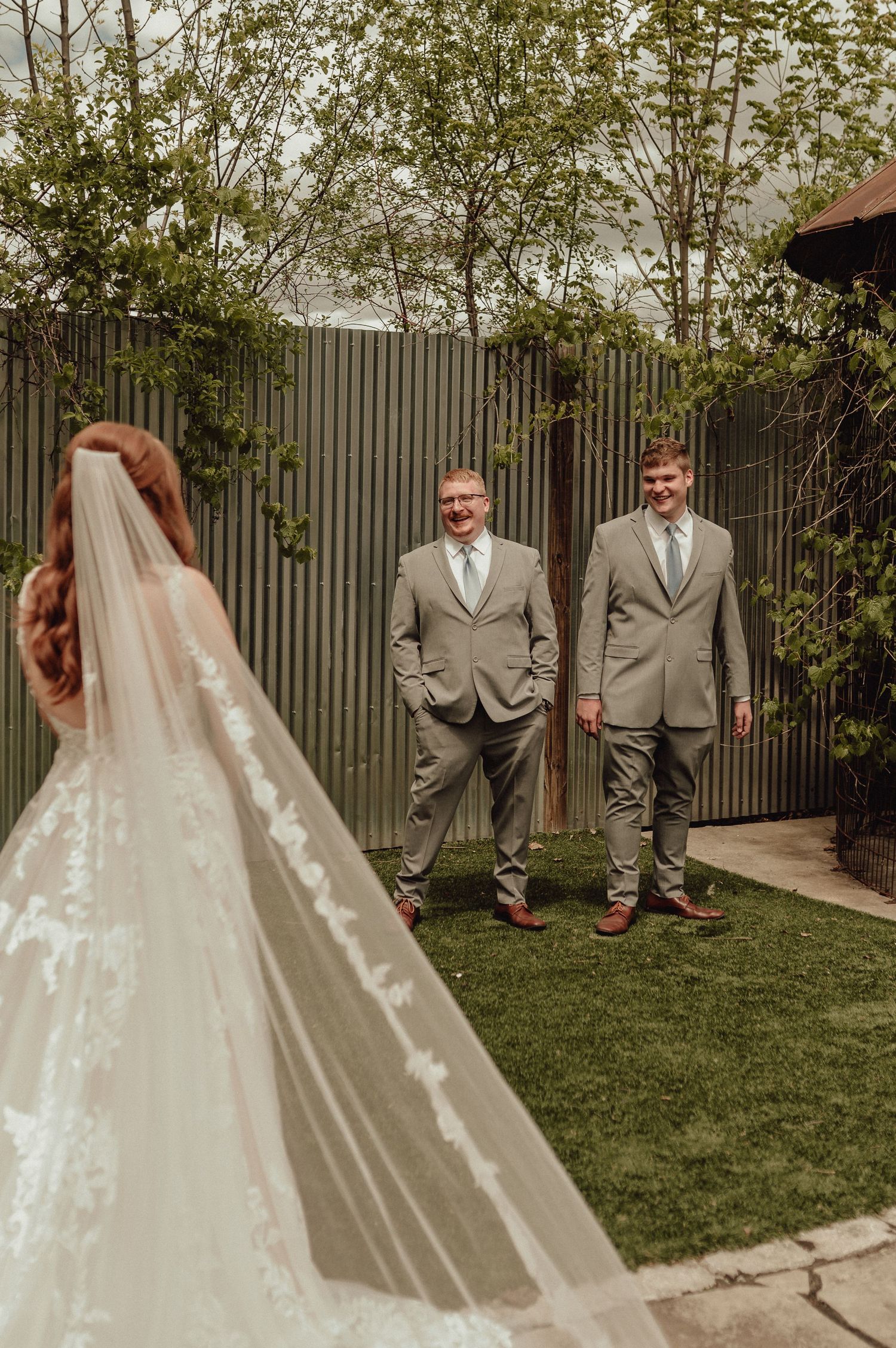 Two groomsmen in light gray suits stand on grass while admiring a bride's flowing lace veil at an outdoor wedding.