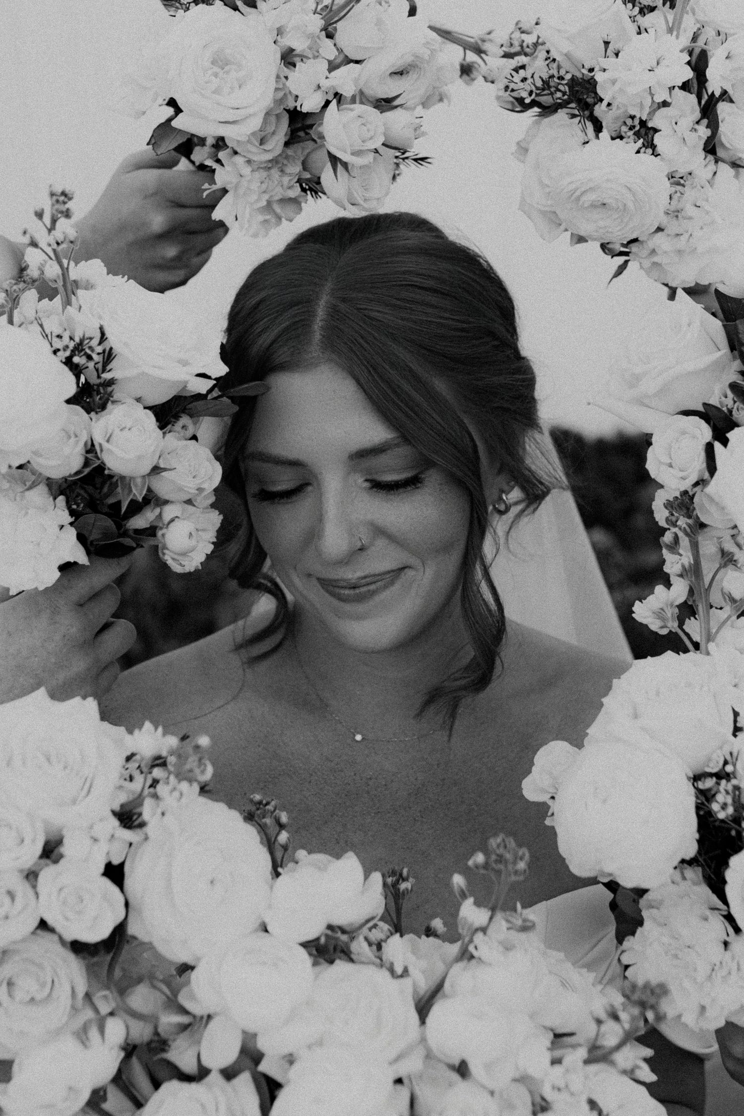 Black and white portrait of a bride surrounded by delicate white roses and flowers in a romantic composition.