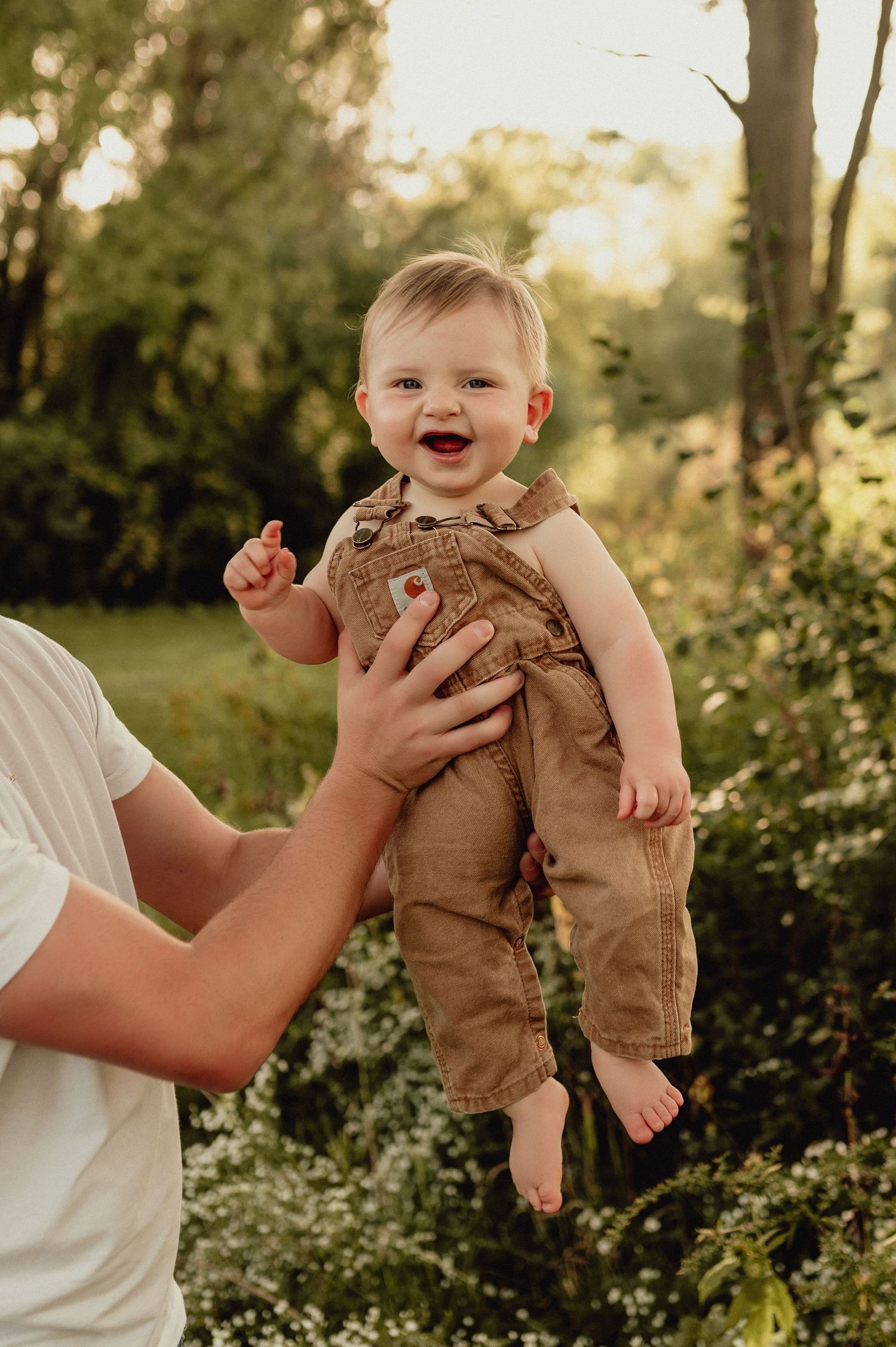 Baby in brown overalls being lifted playfully in garden setting at sunset.
