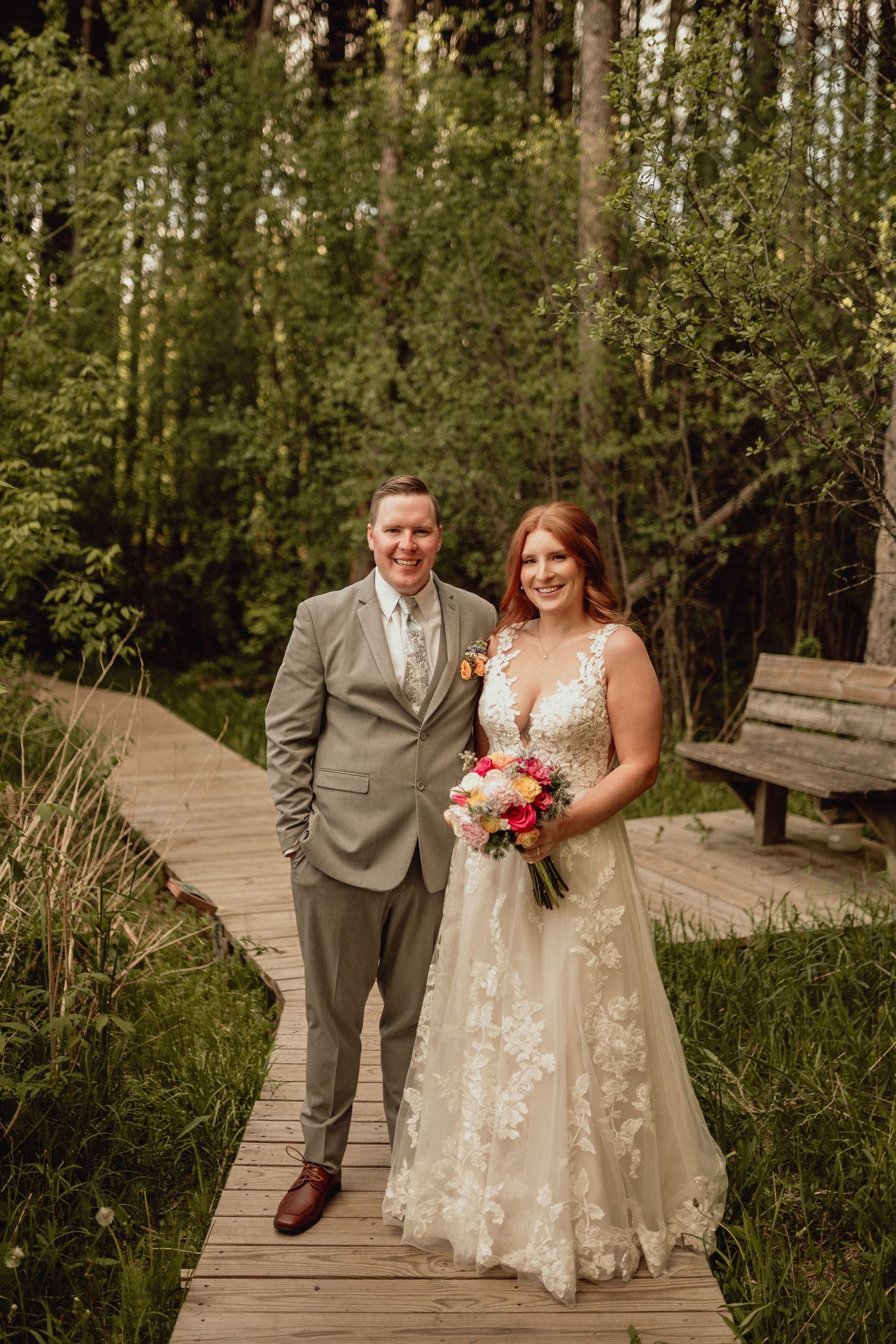 A couple stands together on a wooden boardwalk surrounded by lush green forest during their outdoor wedding.