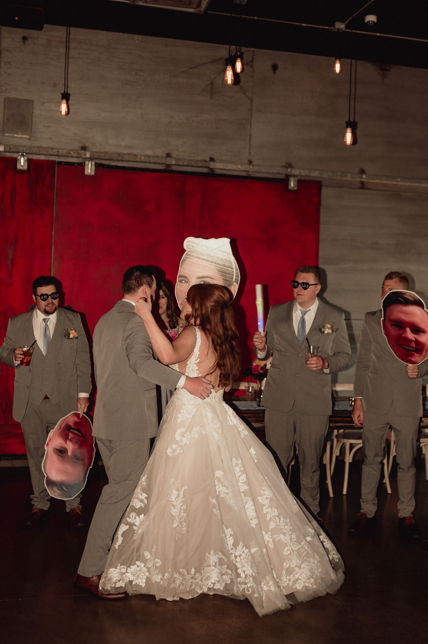 Wedding party in grey suits dances with bride in flowing white lace gown at indoor reception venue with red walls.