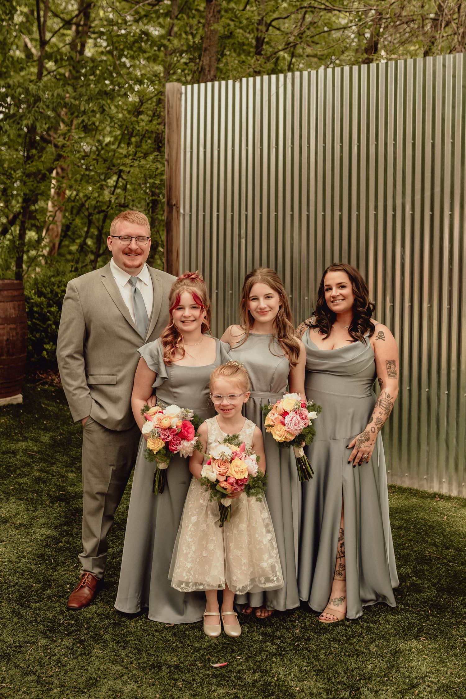 Wedding party poses outdoors in gray and white attire holding pink floral bouquets against a metal fence.