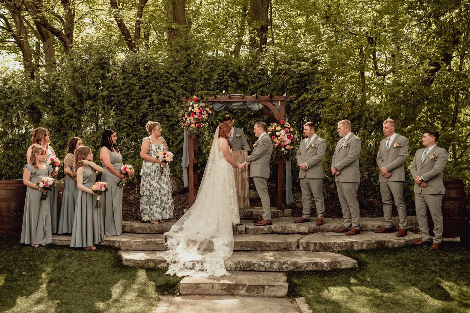 Wedding party stands in line outdoors under trees during summer ceremony.