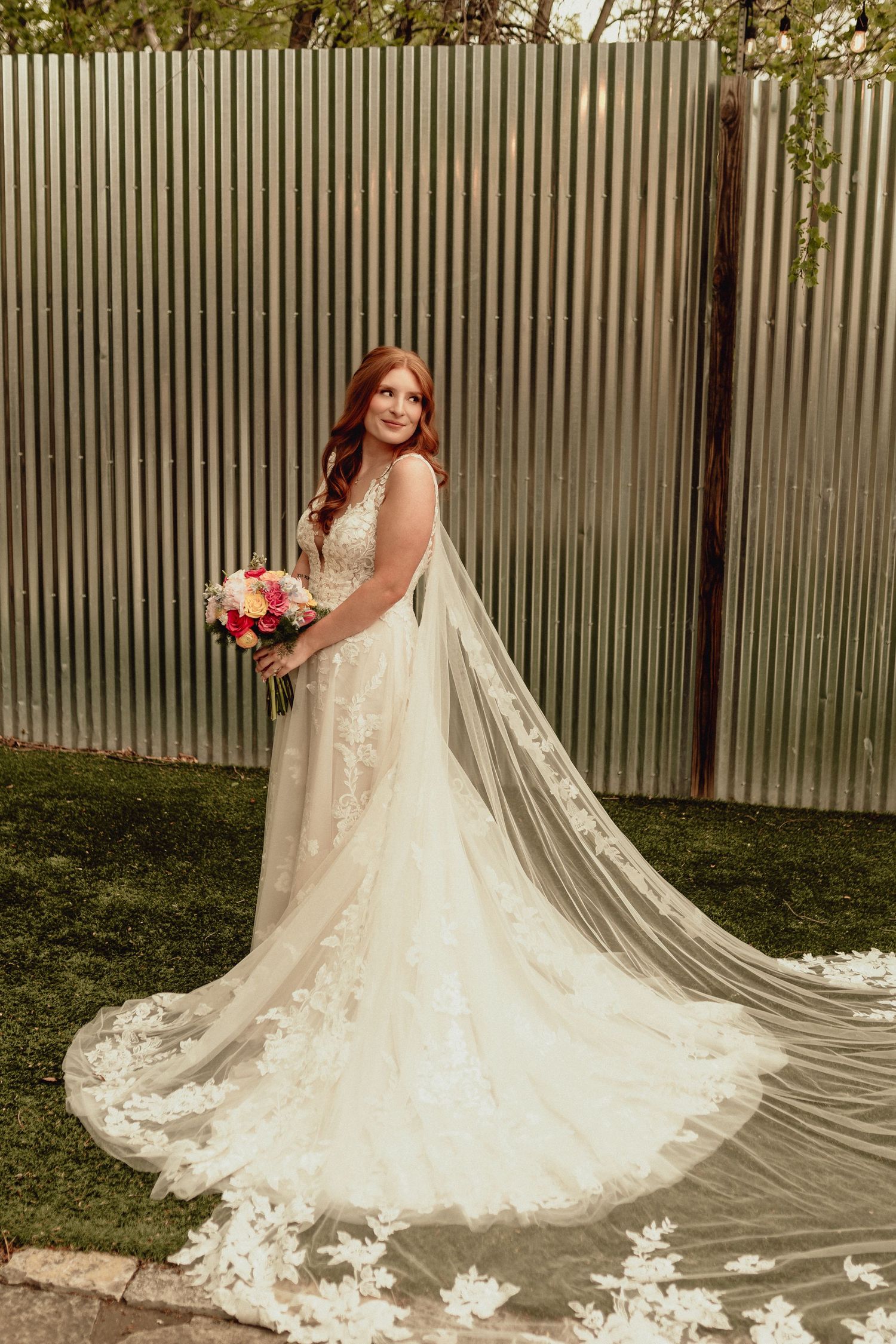 A bride in a lace wedding gown with a long flowing veil holds a colorful bouquet against a corrugated metal fence.