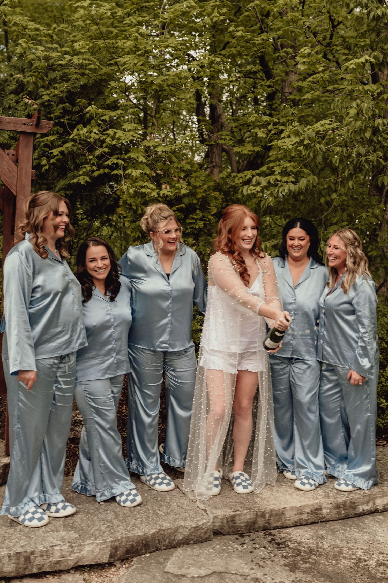 A bridal party wearing matching light blue silk pajamas stands together outdoors while celebrating before the wedding.