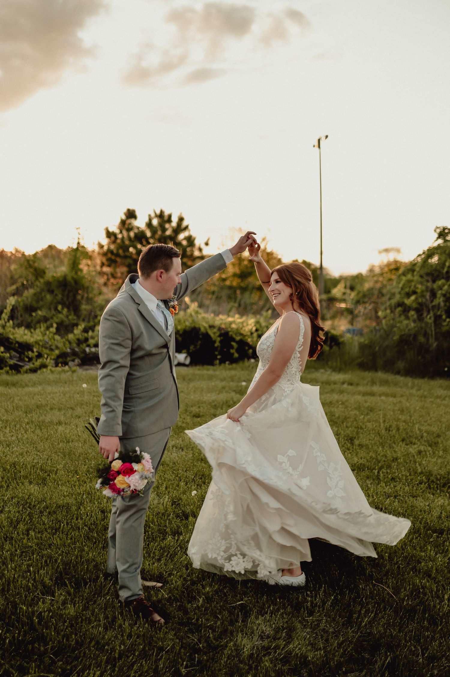 Bride and groom playfully dance together in a grassy field at sunset, with her white dress flowing in the breeze.