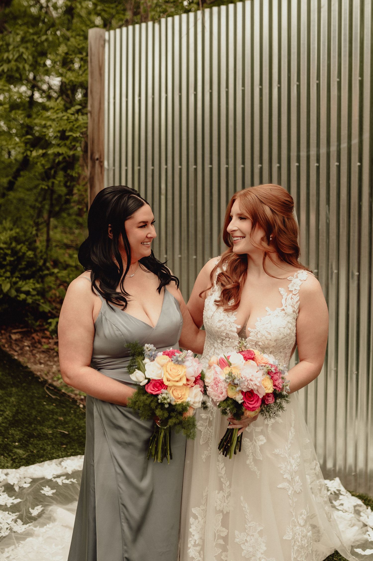 Two women in wedding attire smile at each other while holding beautiful pink and peach floral bouquets.