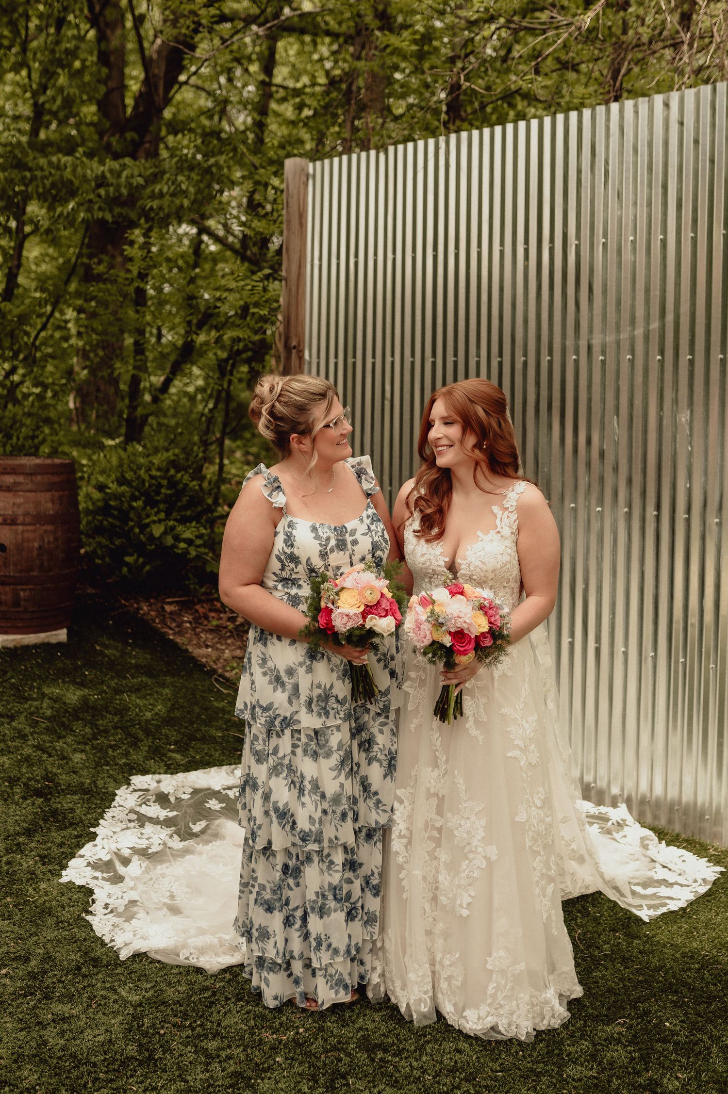 Two brides share a joyful moment outdoors while holding colorful bouquets in front of a white picket fence.