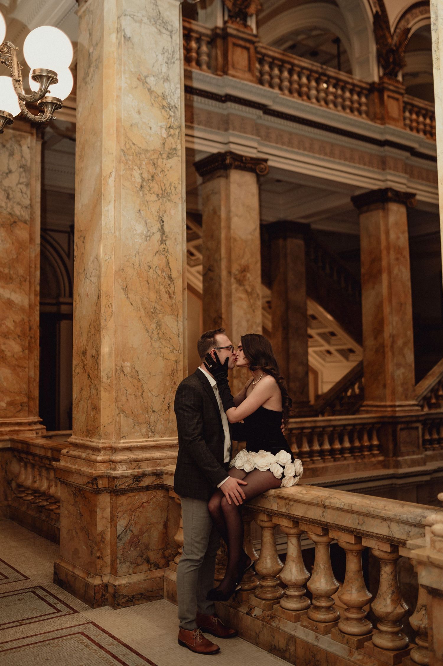 A romantic couple embraces on a grand marble staircase in an ornate historic building with classical architecture.