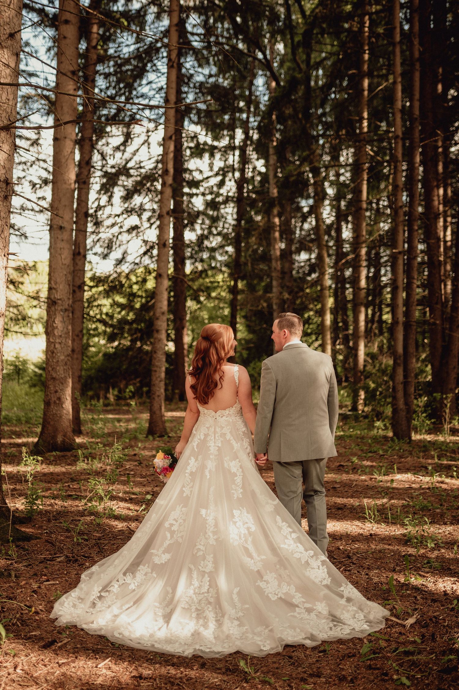 Romantic wedding couple walks through sunlit pine forest with bride wearing flowing lace gown with long train.