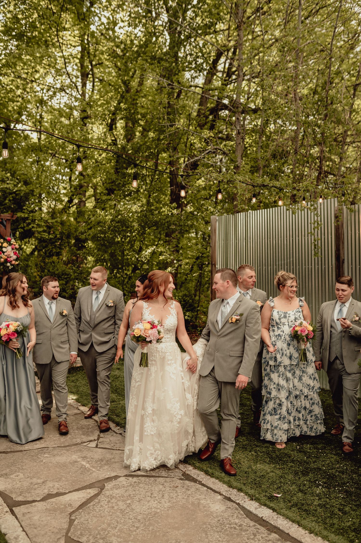 Wedding party in gray and floral attire poses together outdoors along a garden path with greenery in the background.