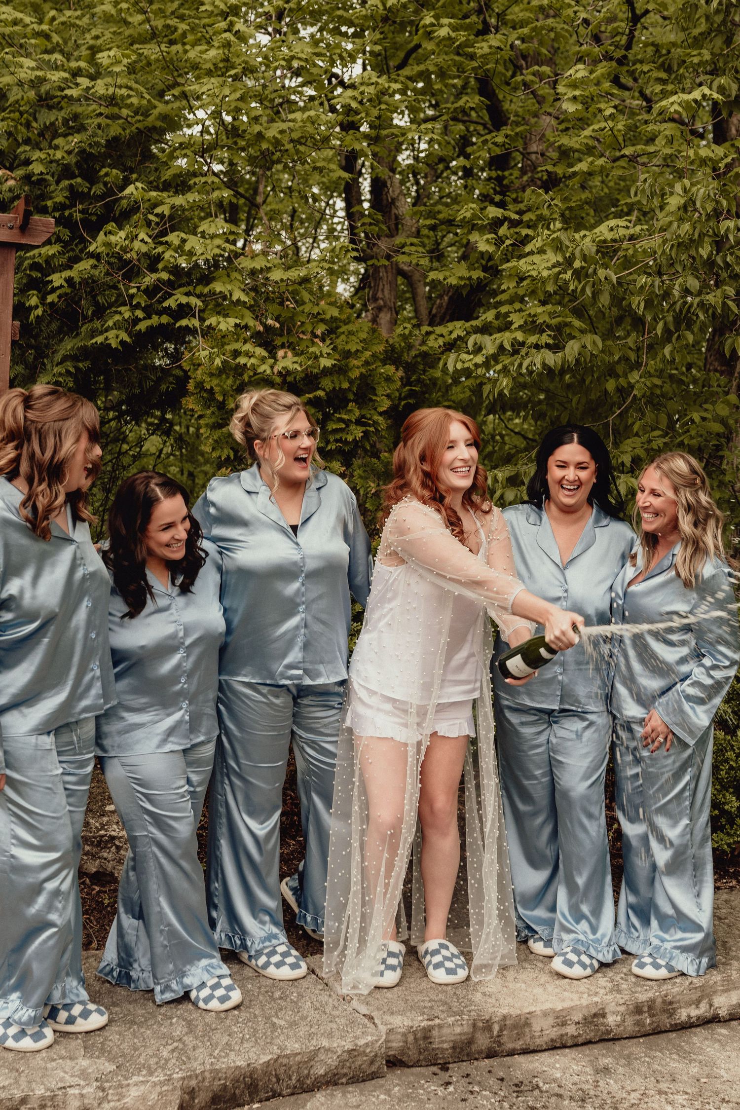 Wedding party wearing light blue suits and a cream dress laugh together while posing on a stone wall outdoors.