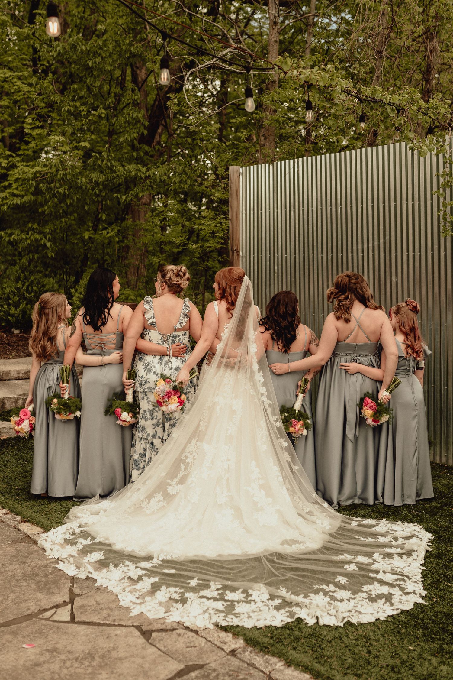 A wedding party in gray dresses and a bride in a lace gown with a flowing train pose together outdoors.