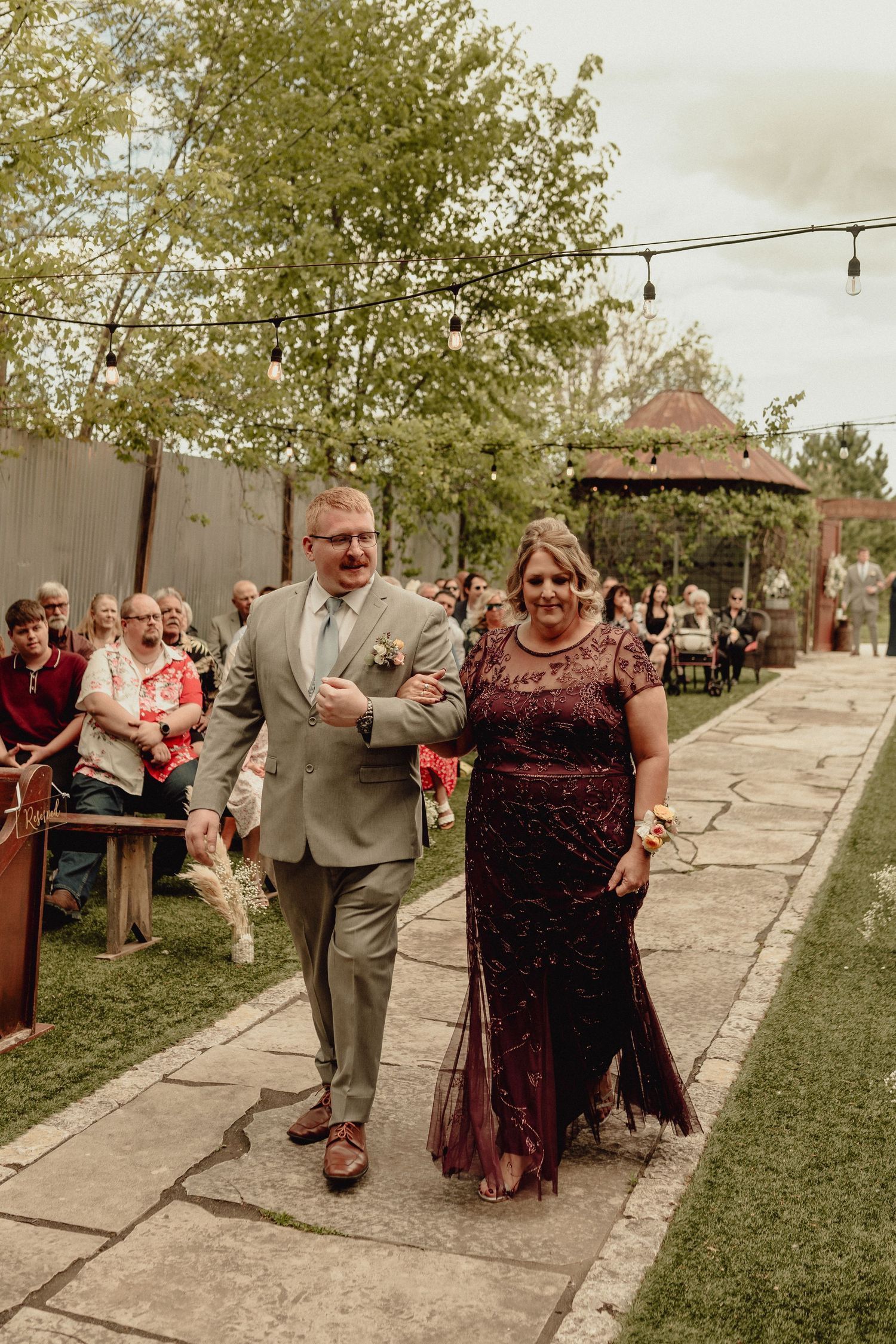 A couple walks down a stone path at an outdoor wedding venue with string lights overhead and guests seated nearby.