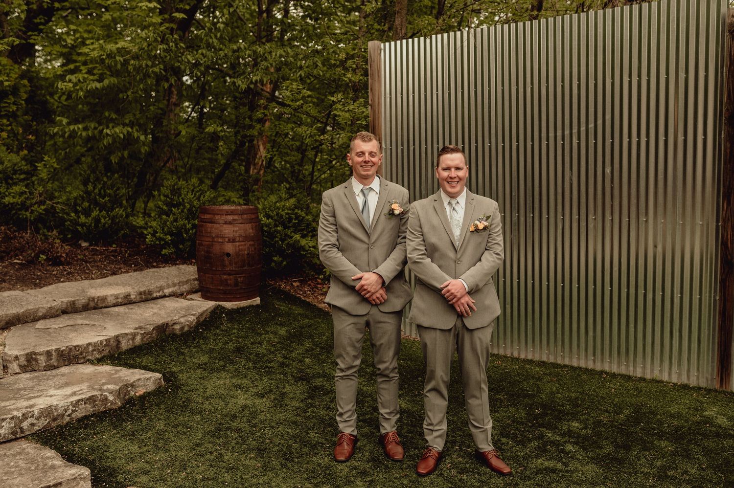 Two people in matching gray suits stand together on stone steps near a metal wall in an outdoor setting.