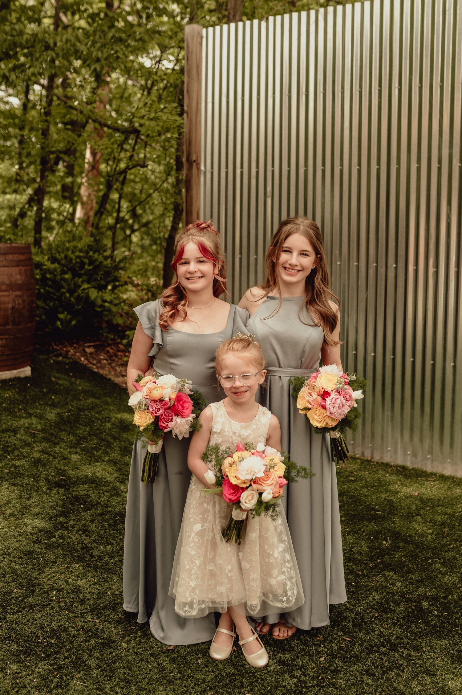 Bridesmaids in gray dresses and flower girl in white pose with colorful bouquets against a metal wall backdrop.