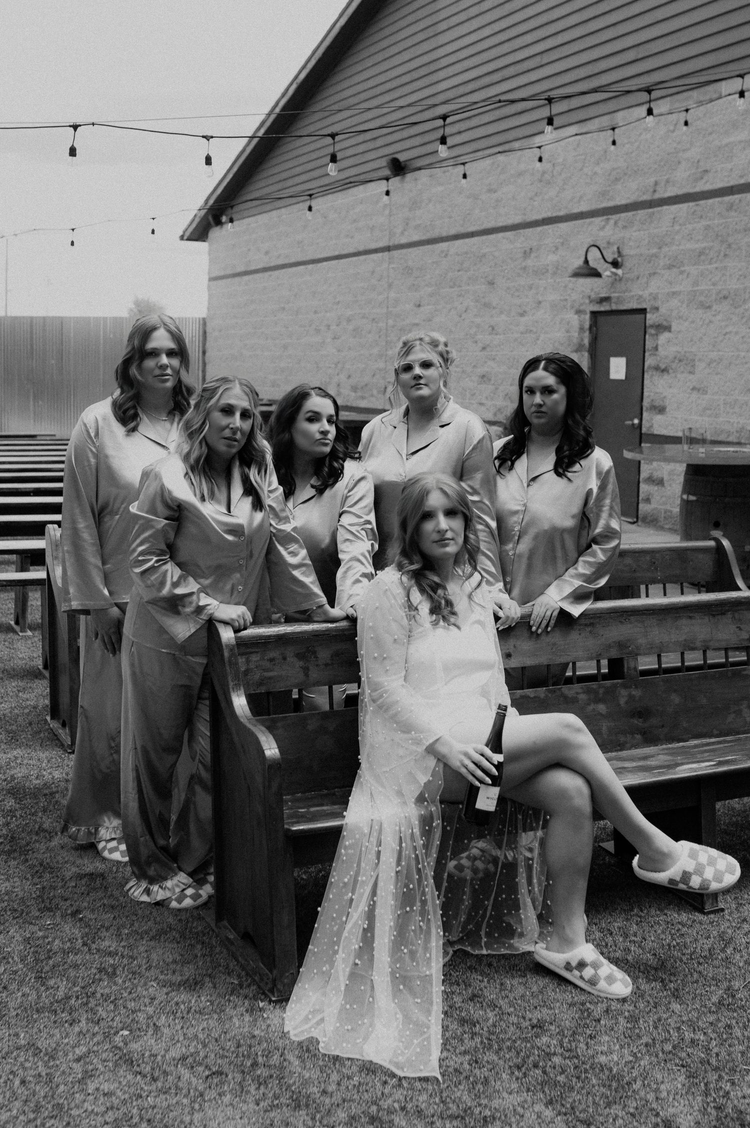 A black and white photograph showing a group of people posing casually on wooden benches outside a building with string lights.