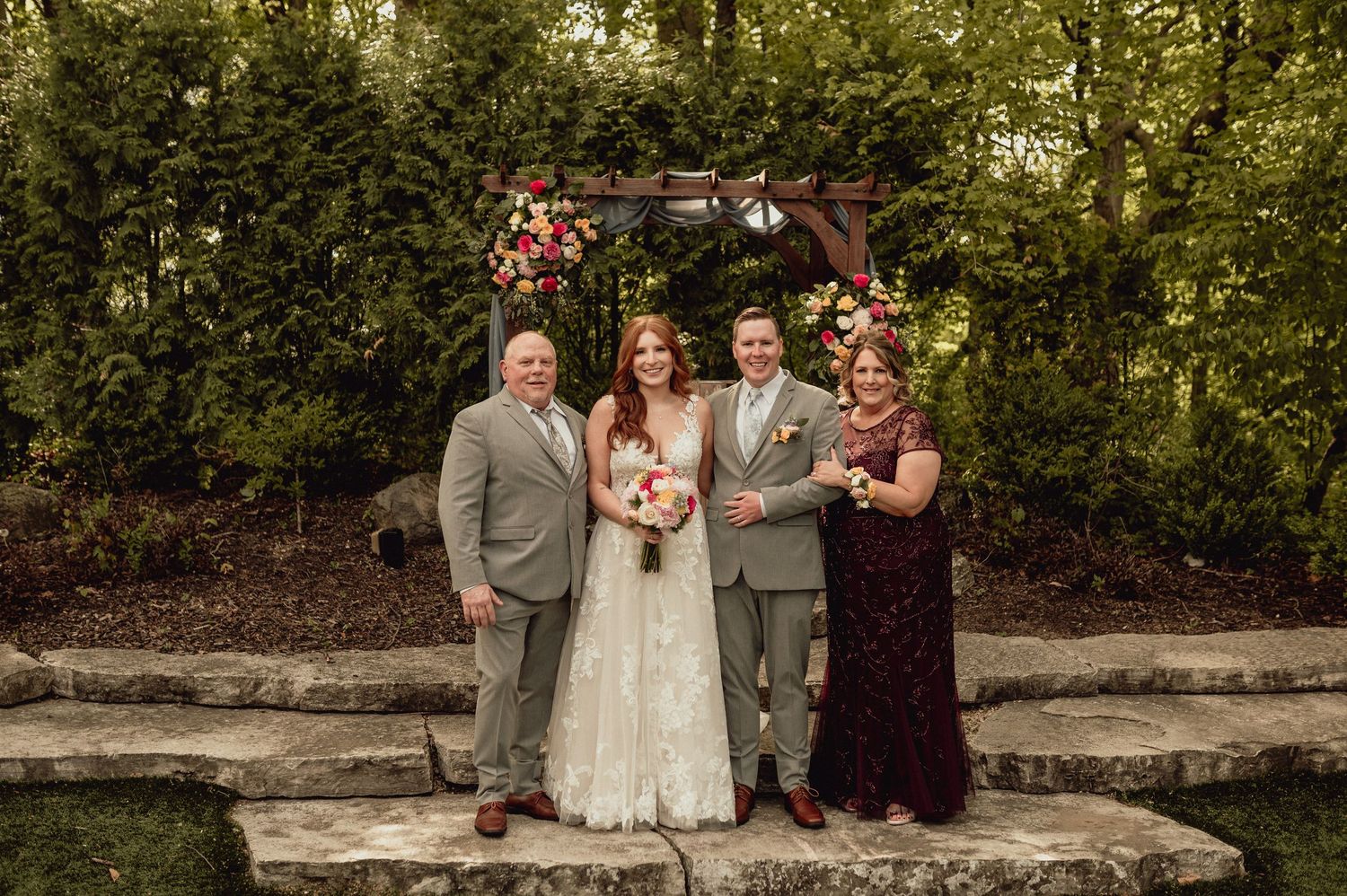 A wedding party poses together on a rustic stone wall surrounded by lush greenery in an outdoor garden setting.