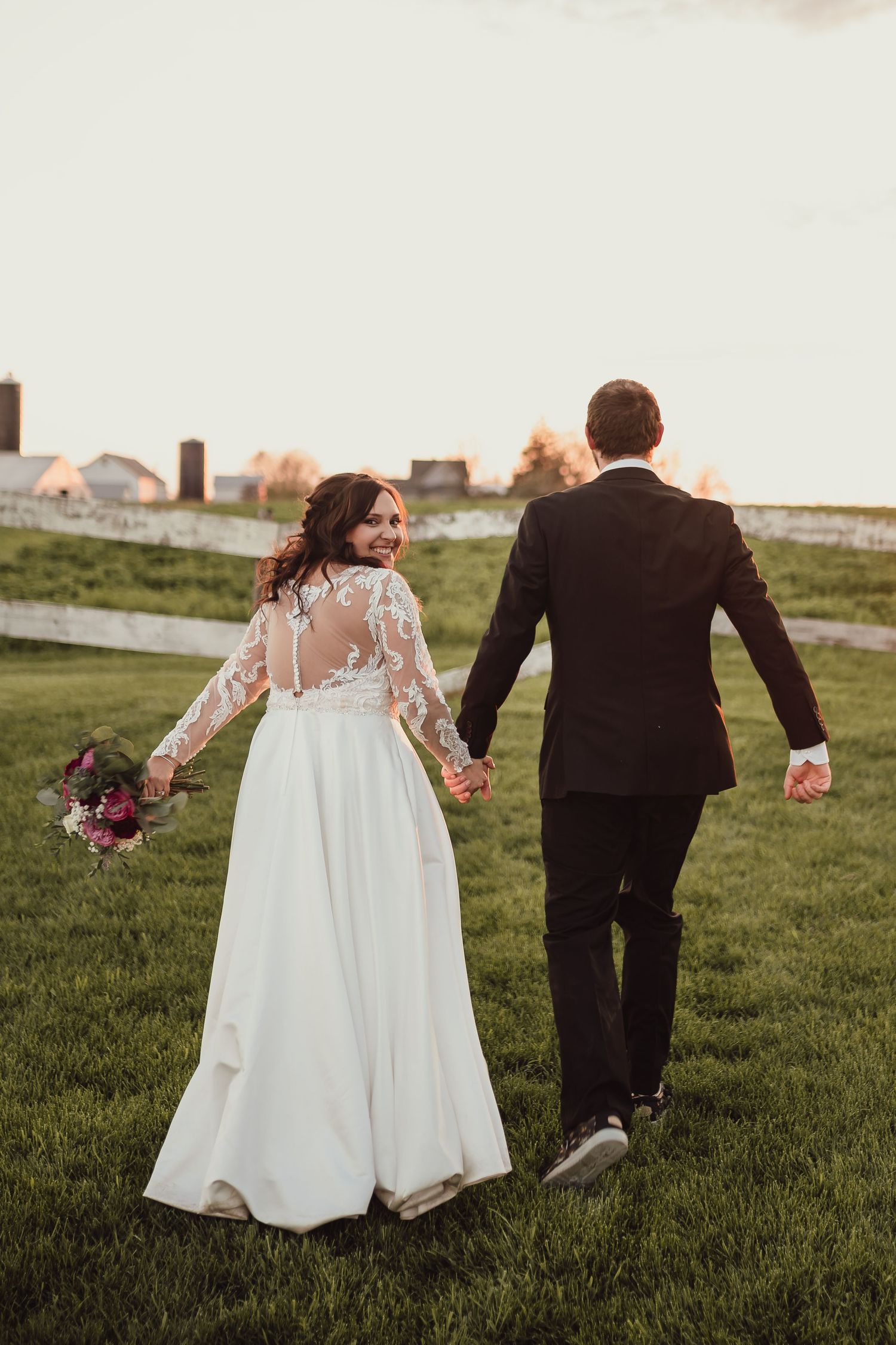 Newlyweds walking hand in hand across green lawn during sunset.