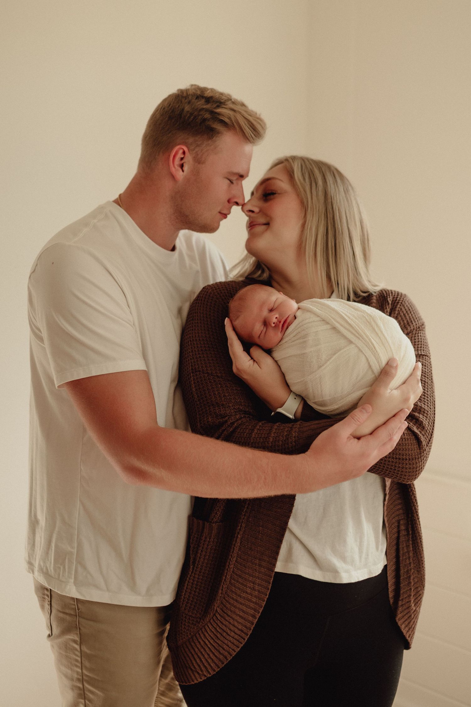 A couple in casual attire sharing an intimate moment while holding a newborn baby wrapped in white.
