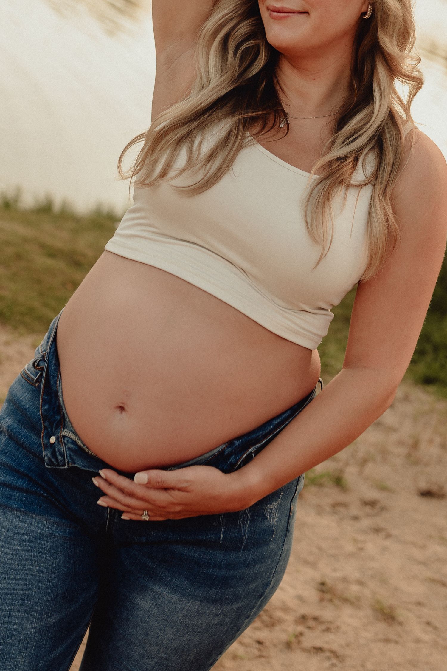 Close-up of pregnant belly in denim jeans and crop top against sandy background.