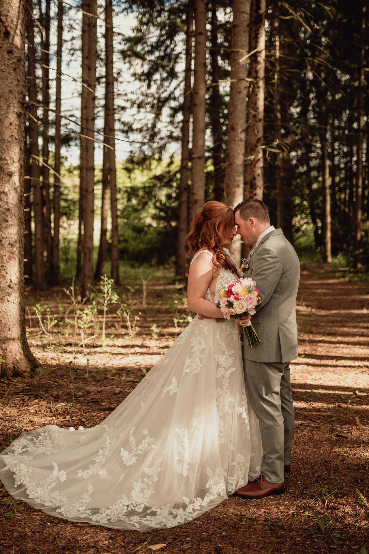 A romantic wedding couple shares an intimate moment in a sunlit pine forest with the bride's lace dress trailing behind them.