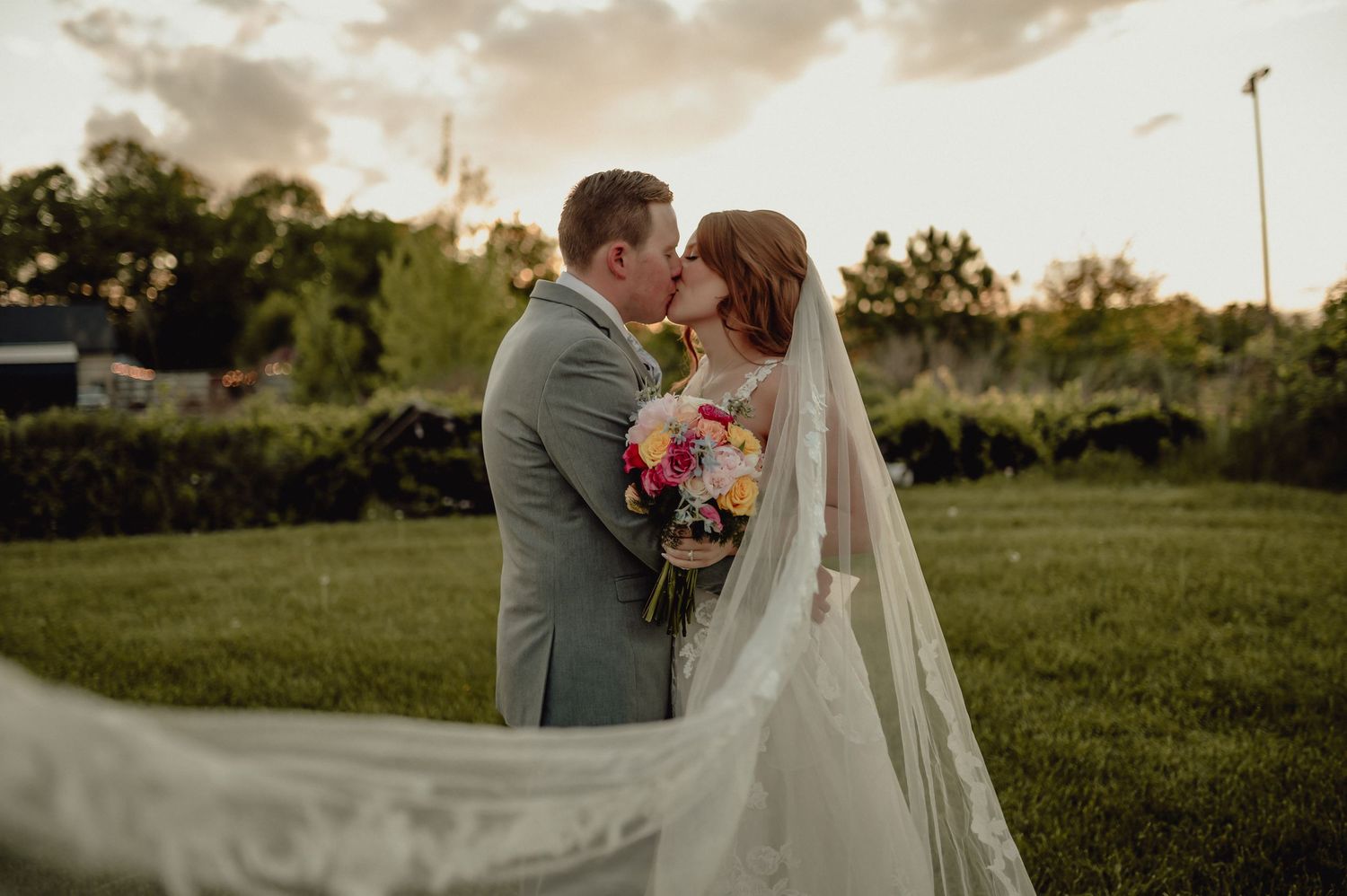 A romantic wedding couple embraces during golden hour in a lush garden setting with a flowing veil in the foreground.