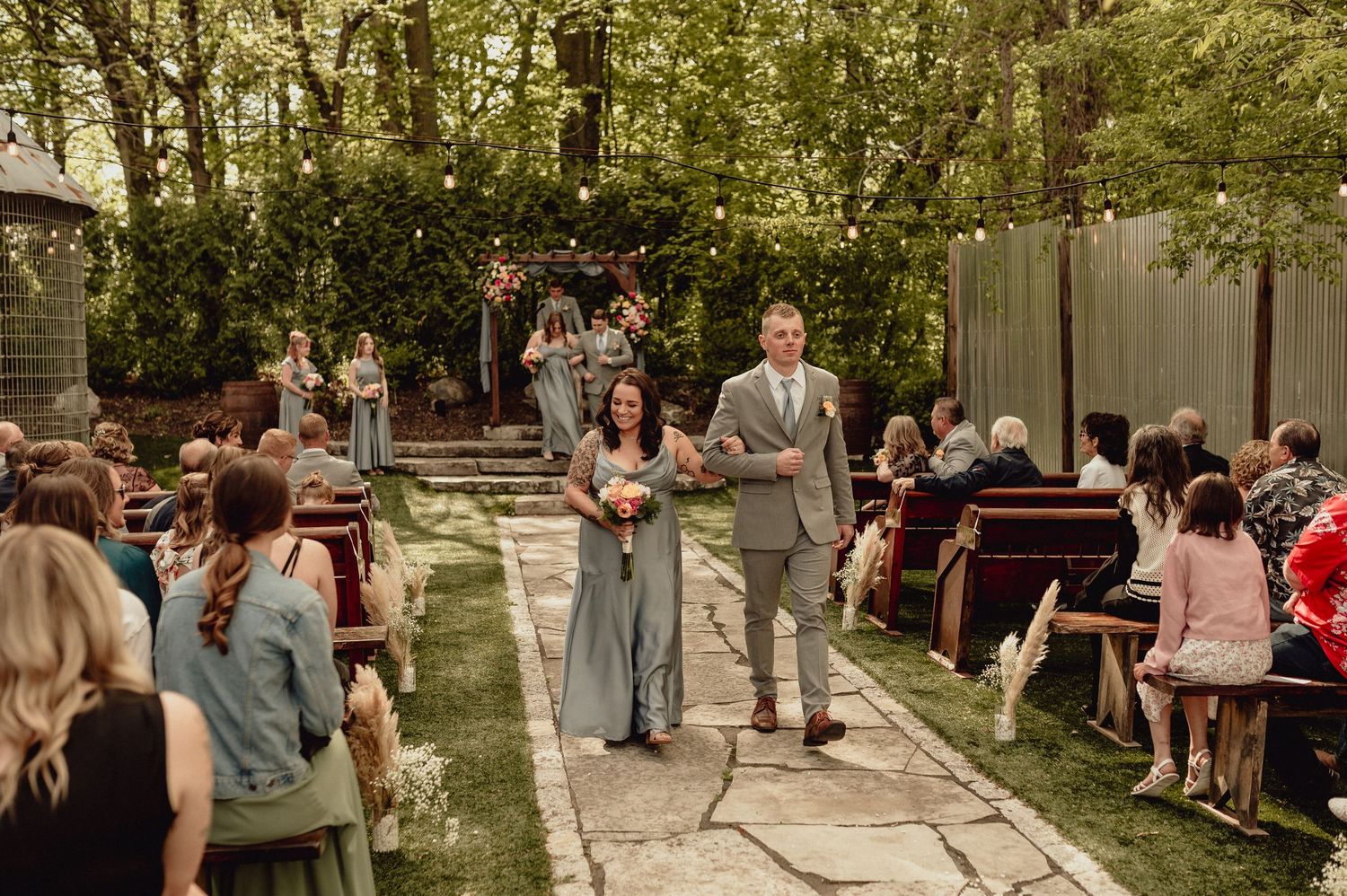 A panoramic view of an outdoor wedding ceremony with guests seated in chairs among trees in a garden setting.