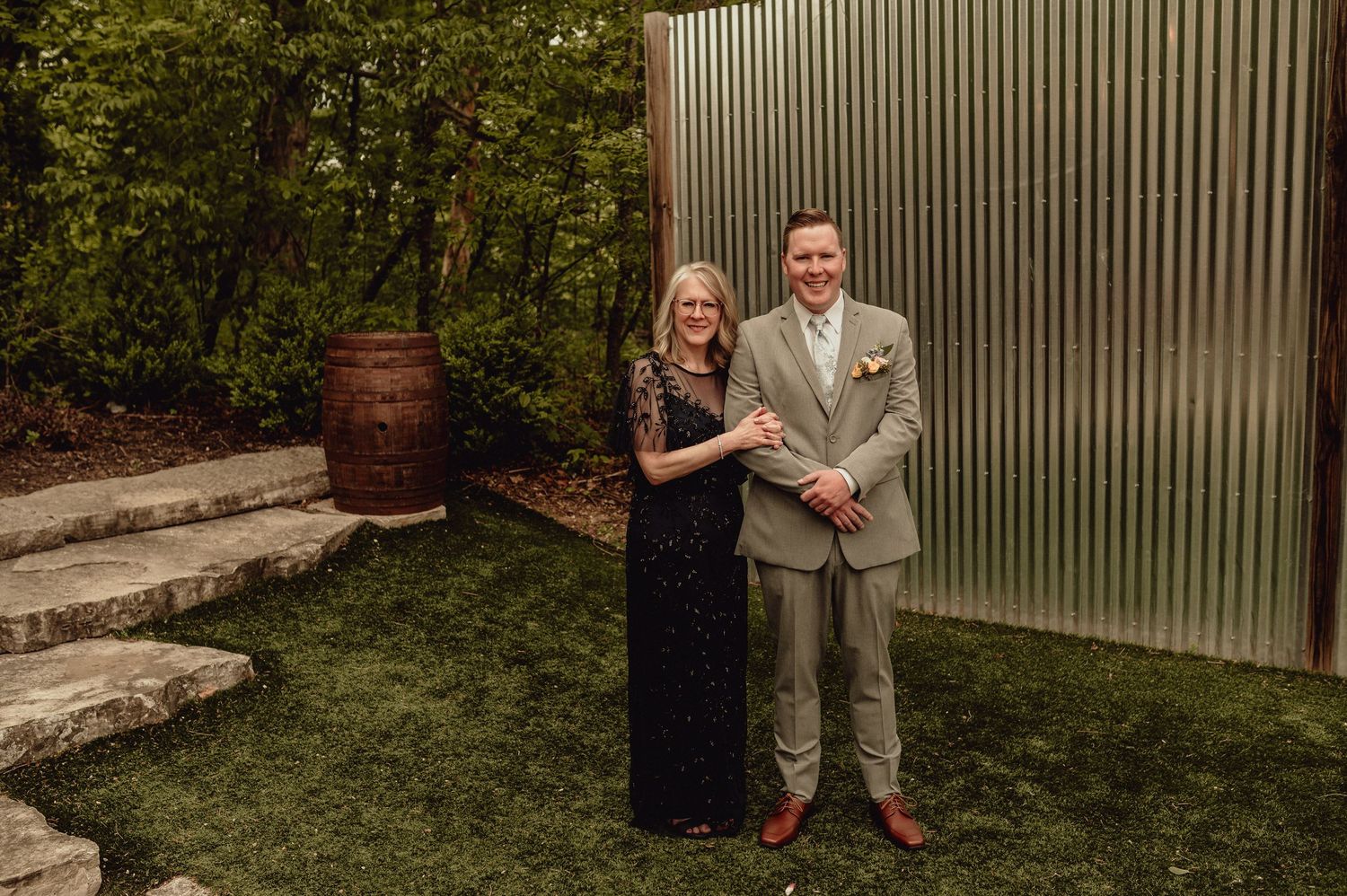 A couple poses together outdoors in formal attire against a metal wall with greenery and a rustic barrel in the background.