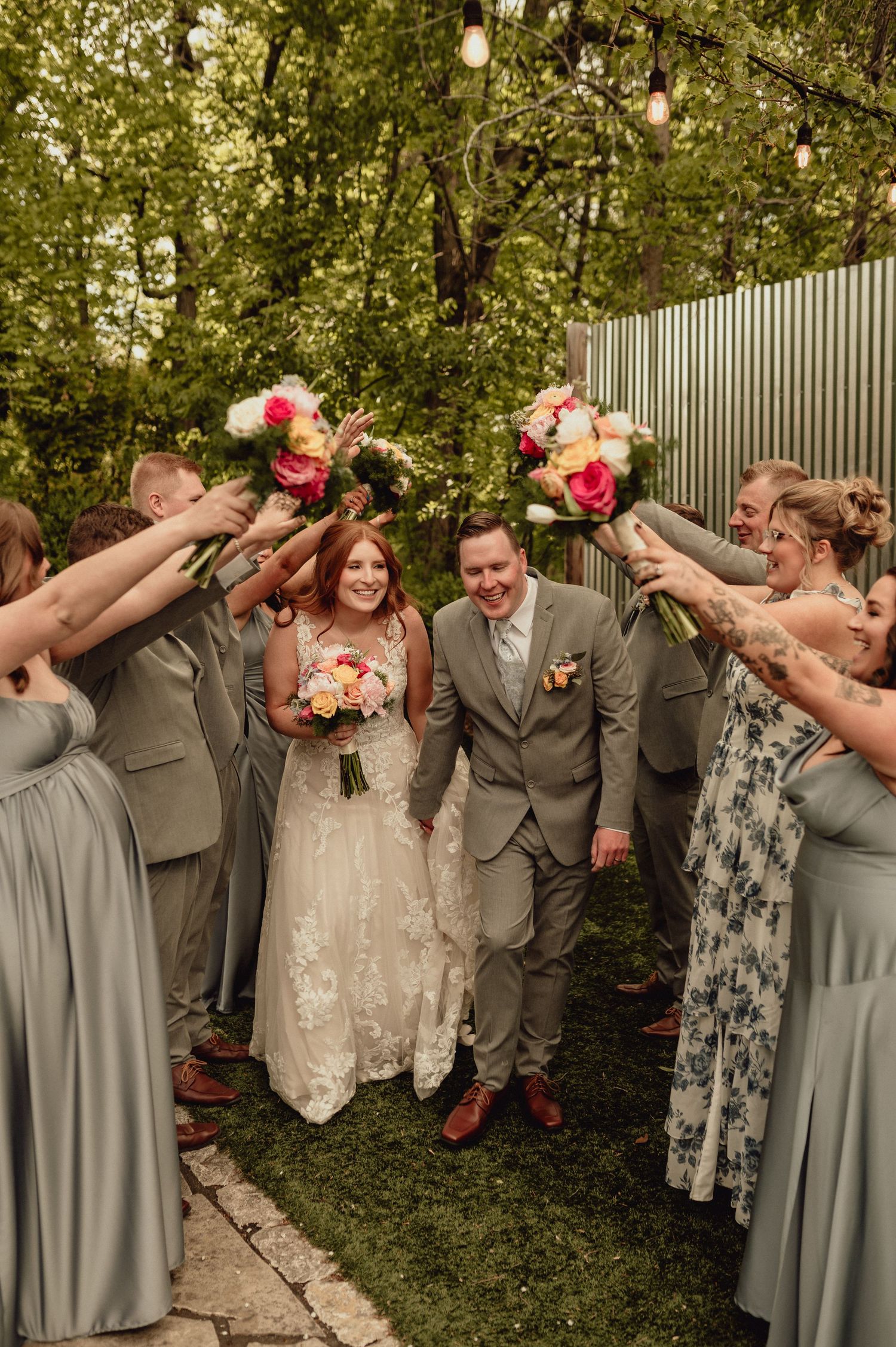 Wedding party in gray and floral attire poses playfully in a garden setting with bouquets of pink and peach flowers.