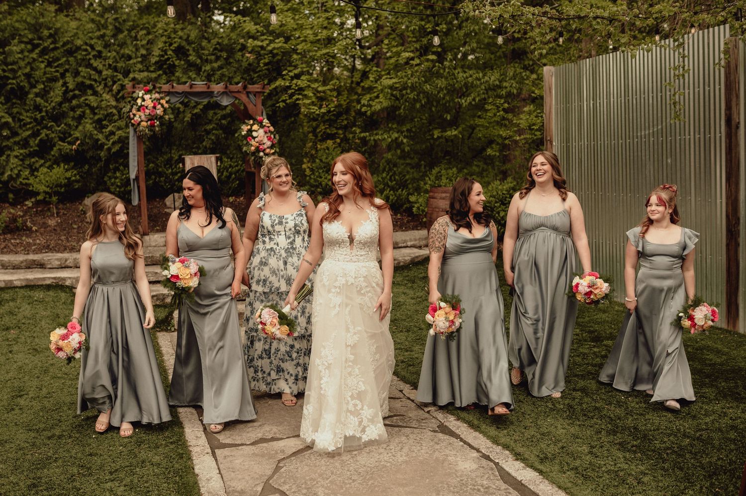 Wedding party in gray bridesmaid dresses and white bridal gown pose together in outdoor garden setting.