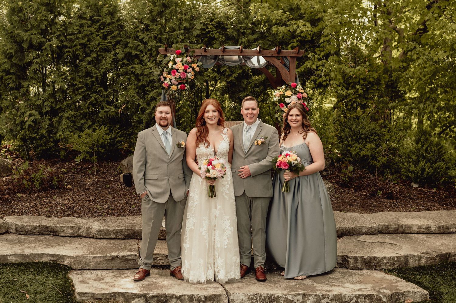 A wedding party poses outdoors in gray and white attire with red floral bouquets against a garden backdrop.