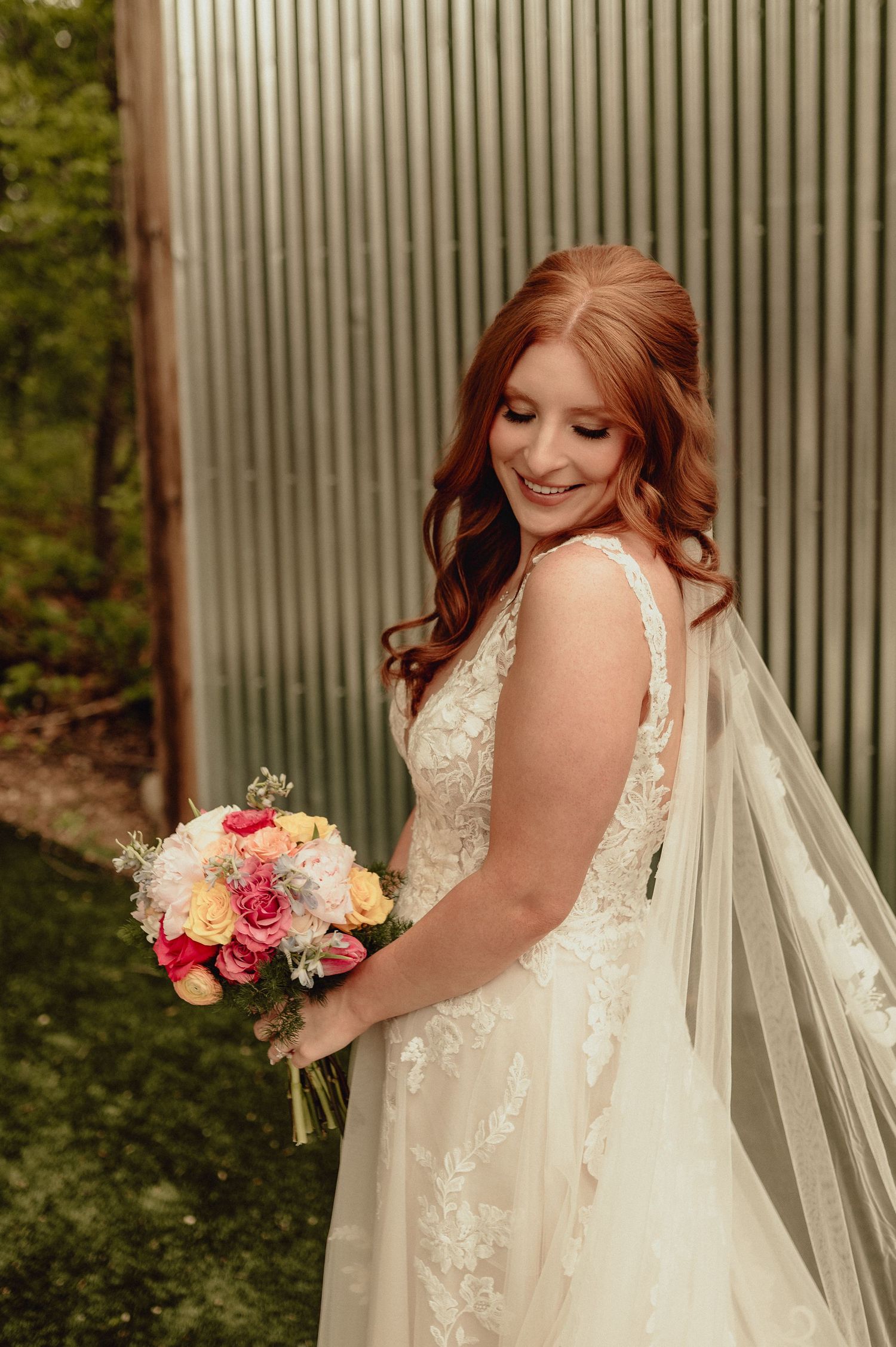 A bride in a lace wedding dress poses with a colorful bouquet against a corrugated metal wall.