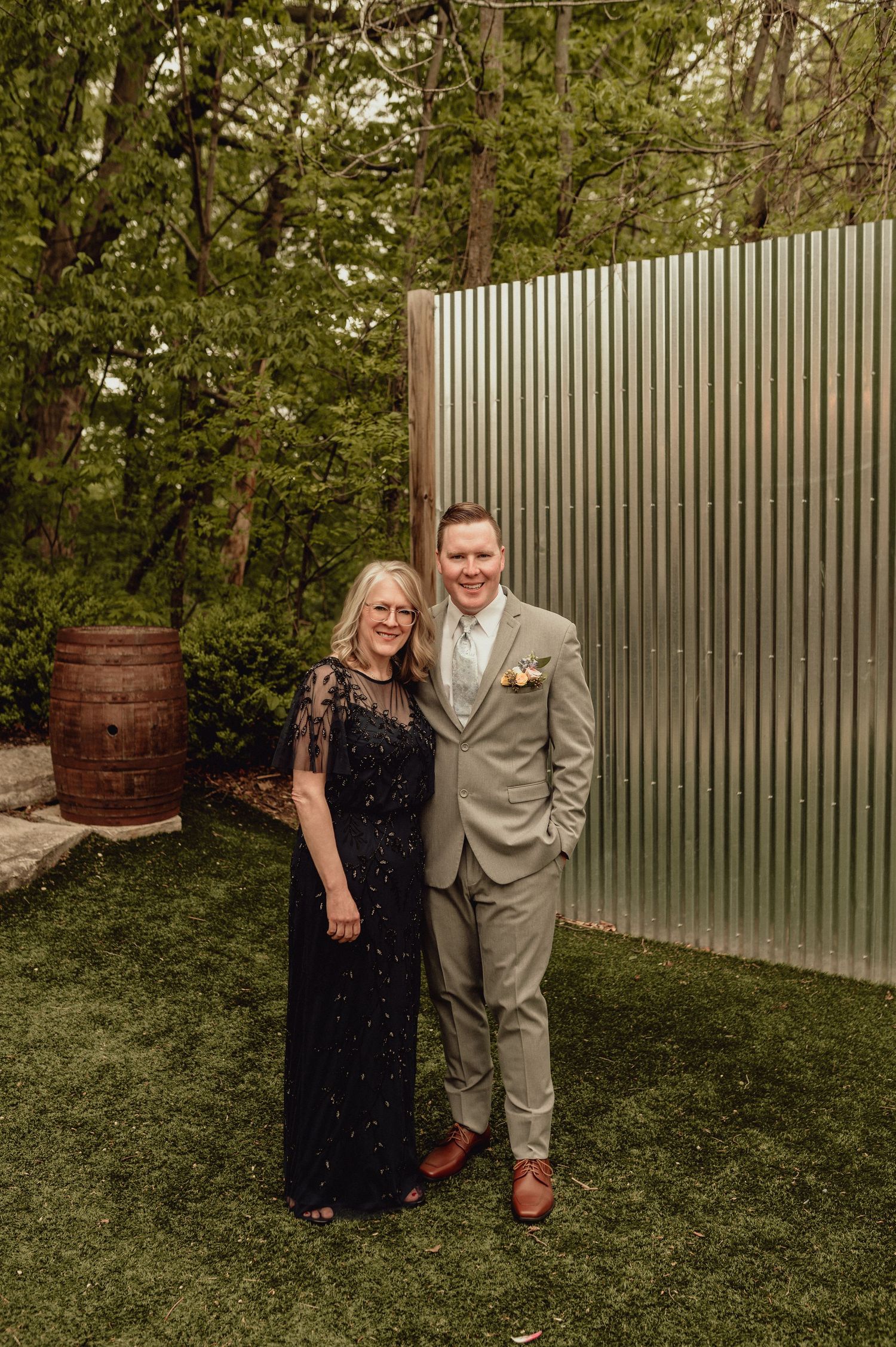 A couple poses together outdoors in formal attire against a backdrop of greenery and a metal fence.