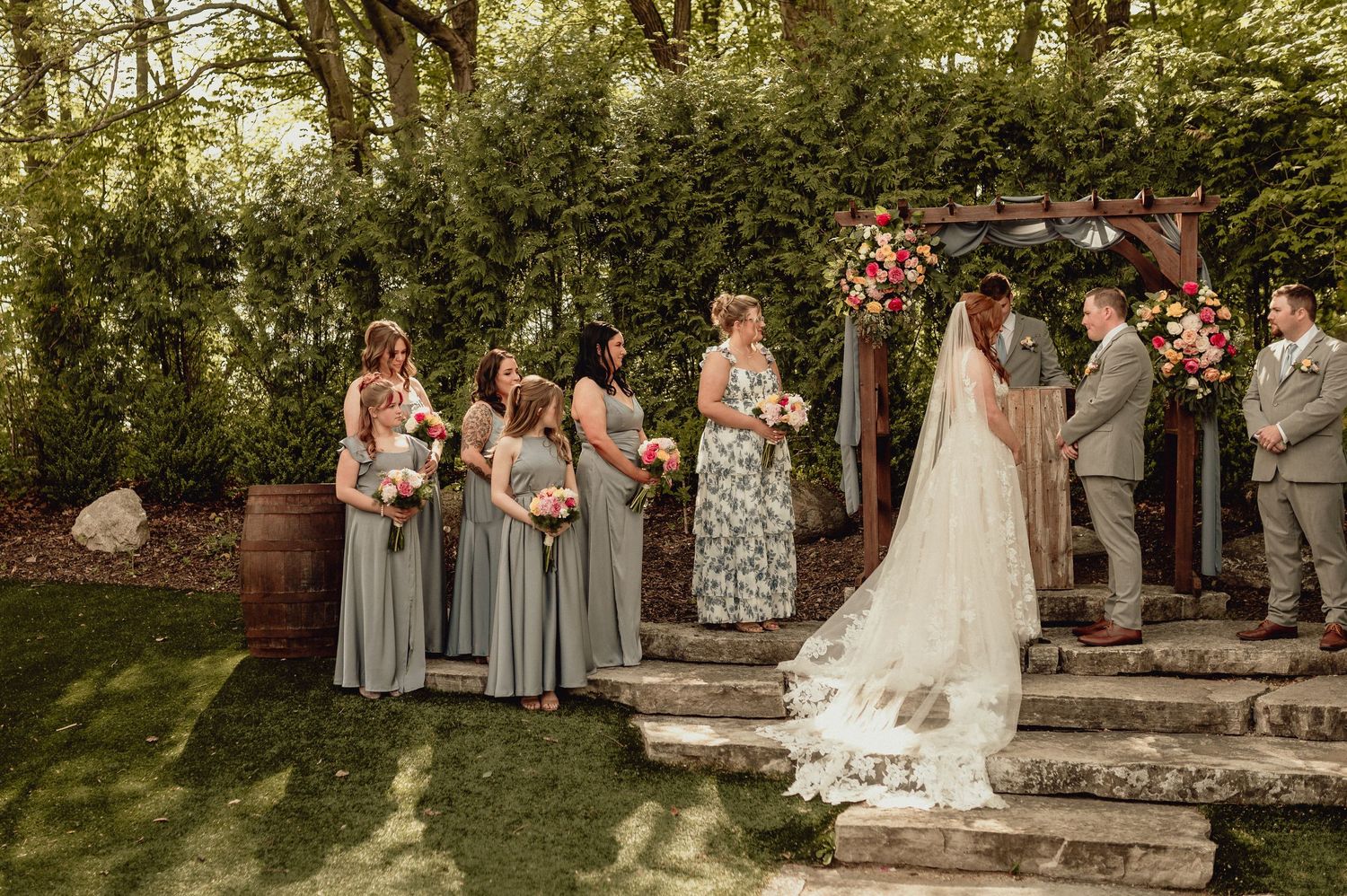 A wedding party stands in a garden setting wearing gray and blush colored attire while posing for formal photos.