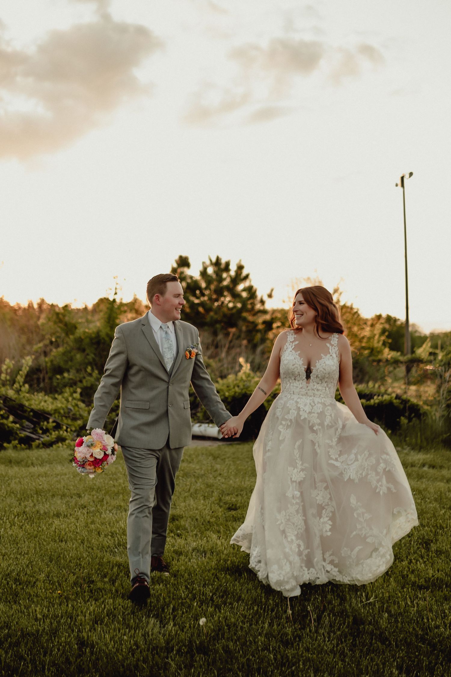 A couple in wedding attire walk hand in hand through a grassy field at sunset during their outdoor ceremony.