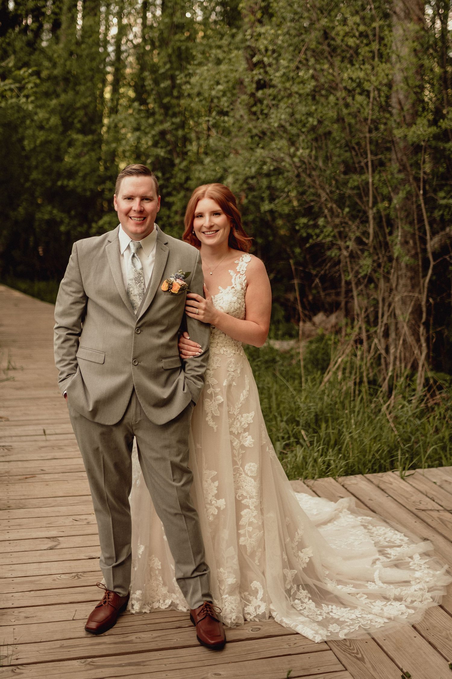 A couple in wedding attire share a joyful moment together on a wooden boardwalk surrounded by lush greenery.