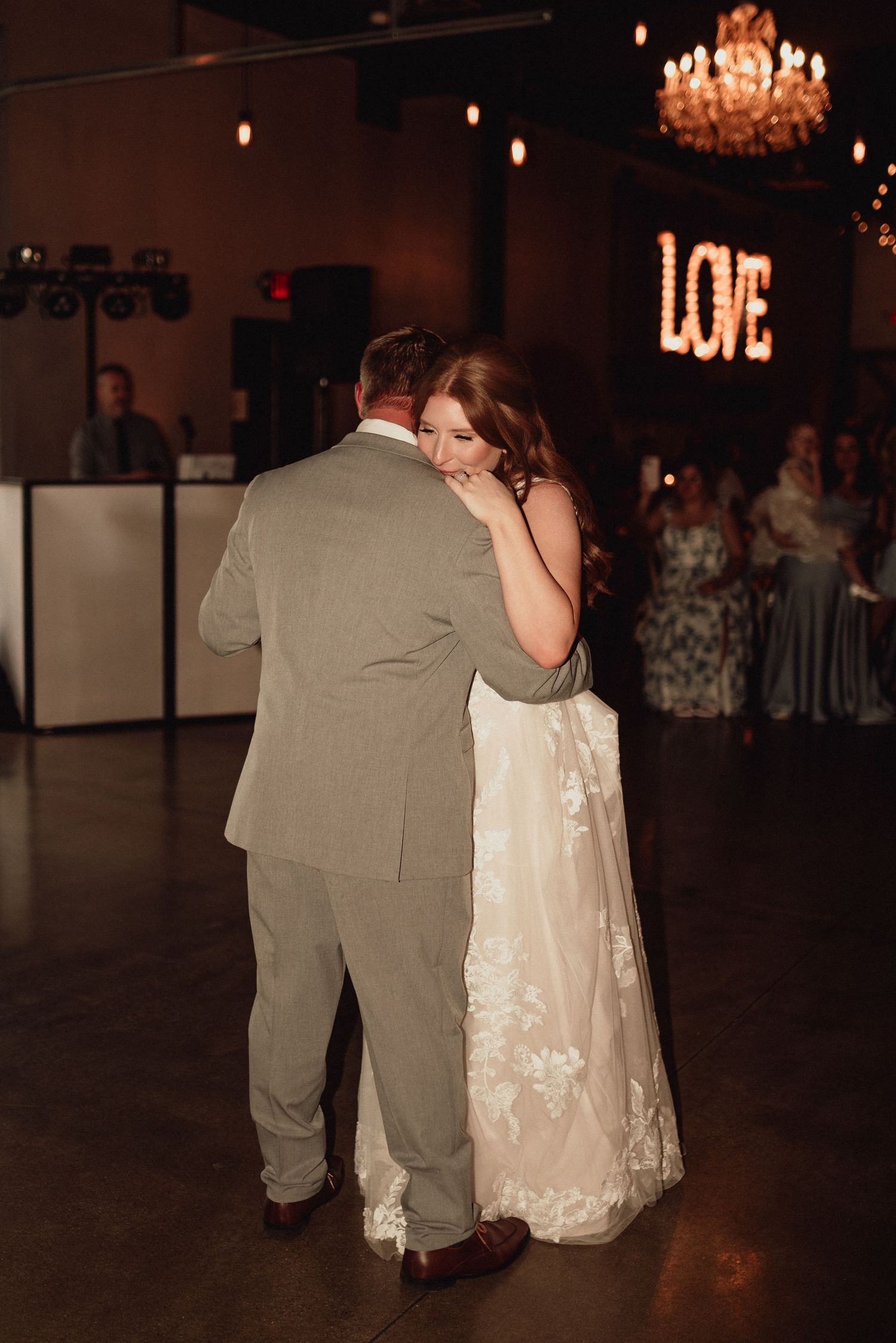 A couple shares their first dance at a wedding reception with illuminated LOVE sign and chandelier in the background.