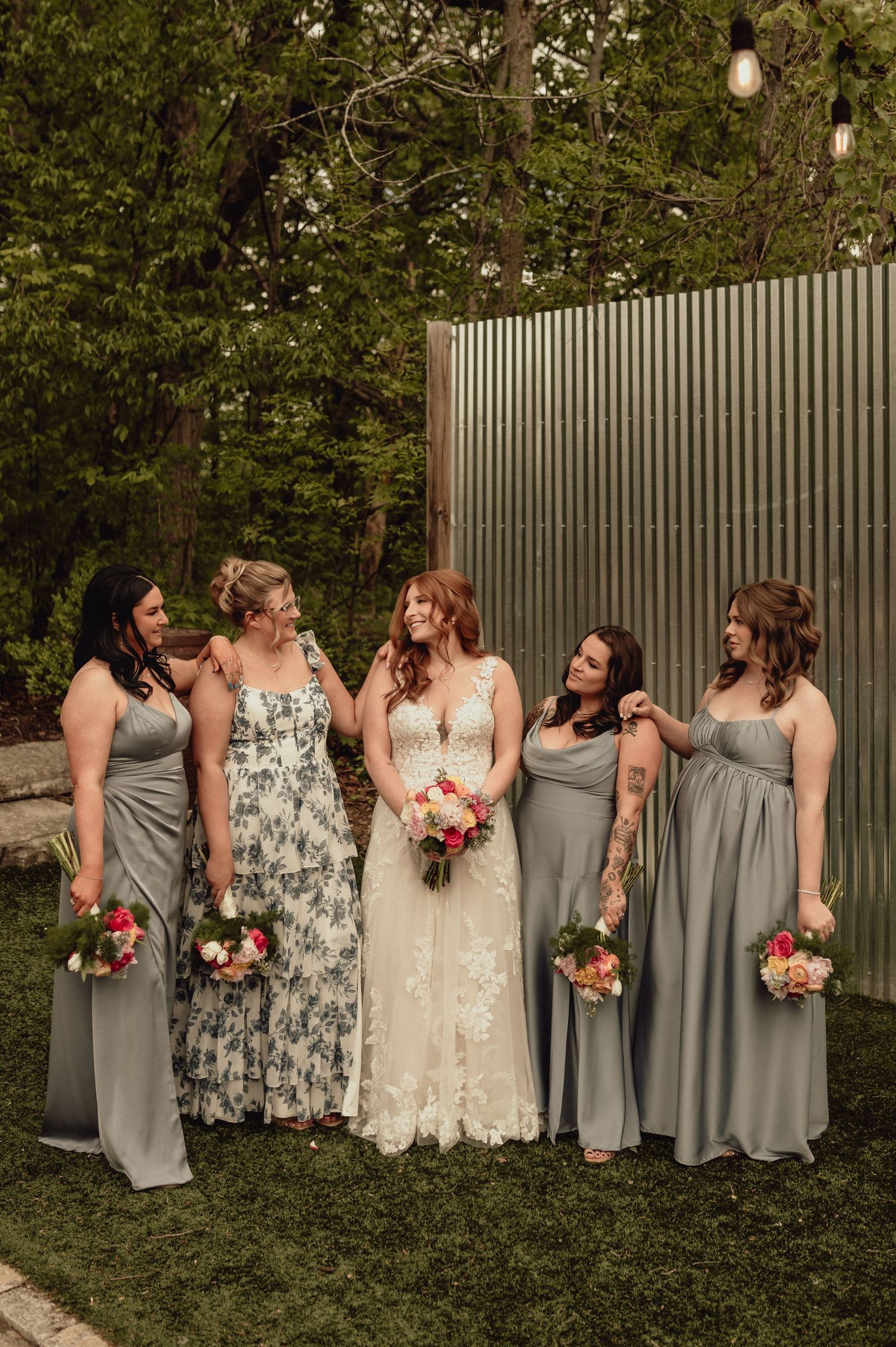 A bridal party poses outdoors in gray bridesmaid dresses and a white lace wedding gown with floral bouquets.
