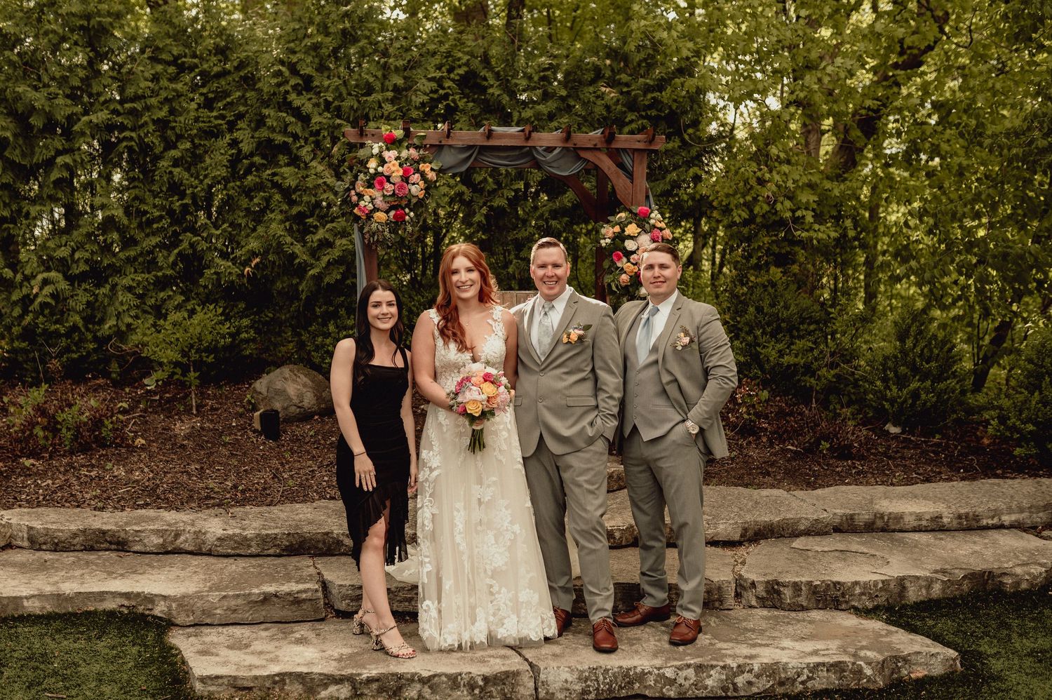Wedding party poses together in front of lush green garden backdrop during outdoor ceremony.