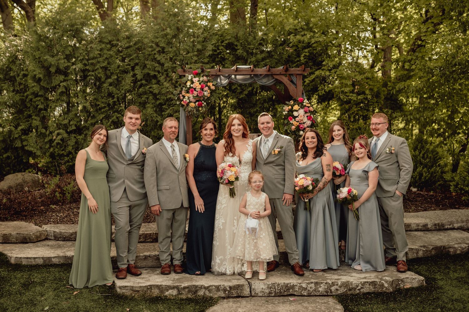 Wedding party members sit and pose together on stone steps surrounded by lush greenery in an outdoor garden setting.