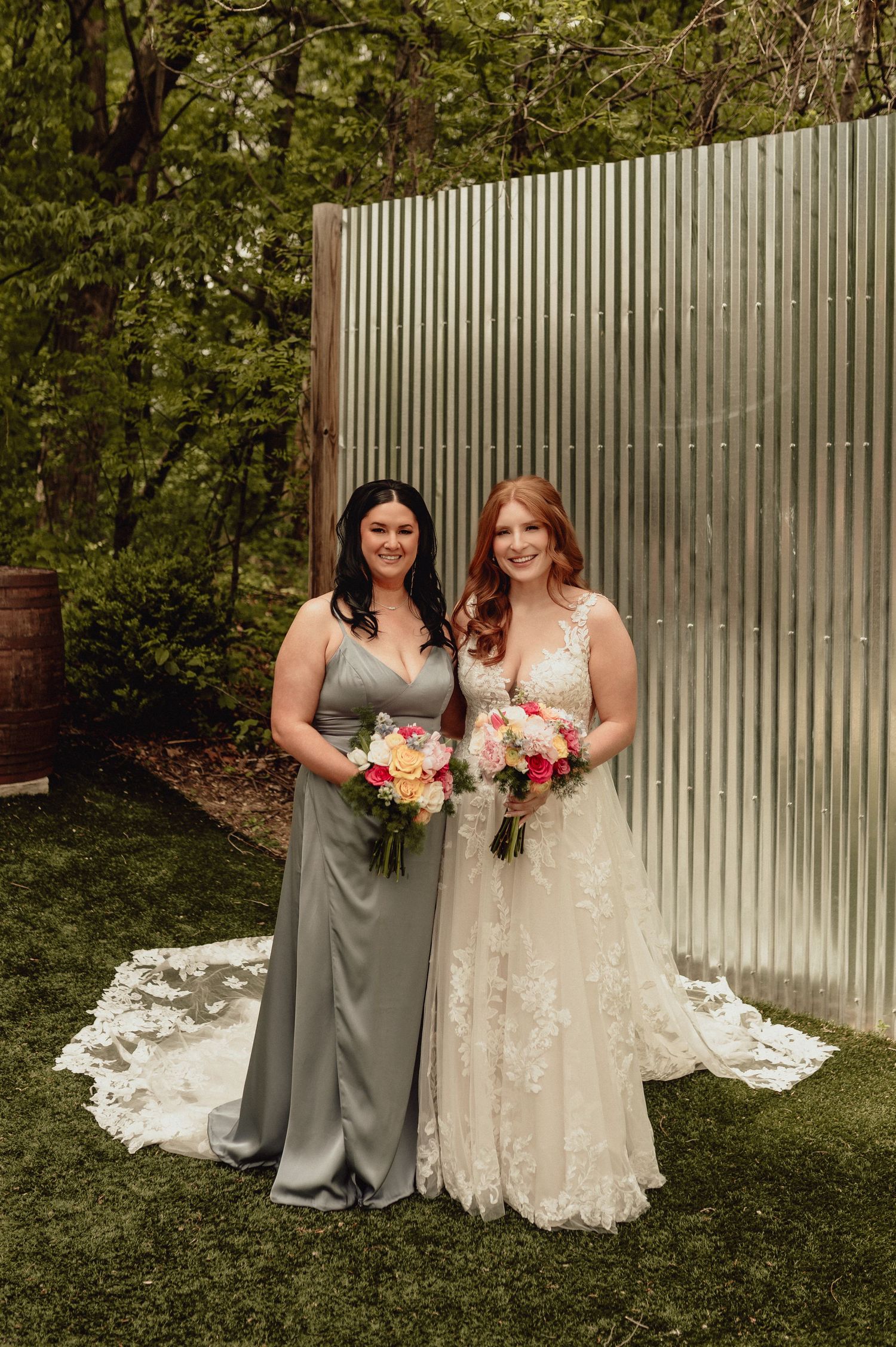 Two people in gray and white dresses pose together with floral bouquets on a grassy lawn near a white fence.