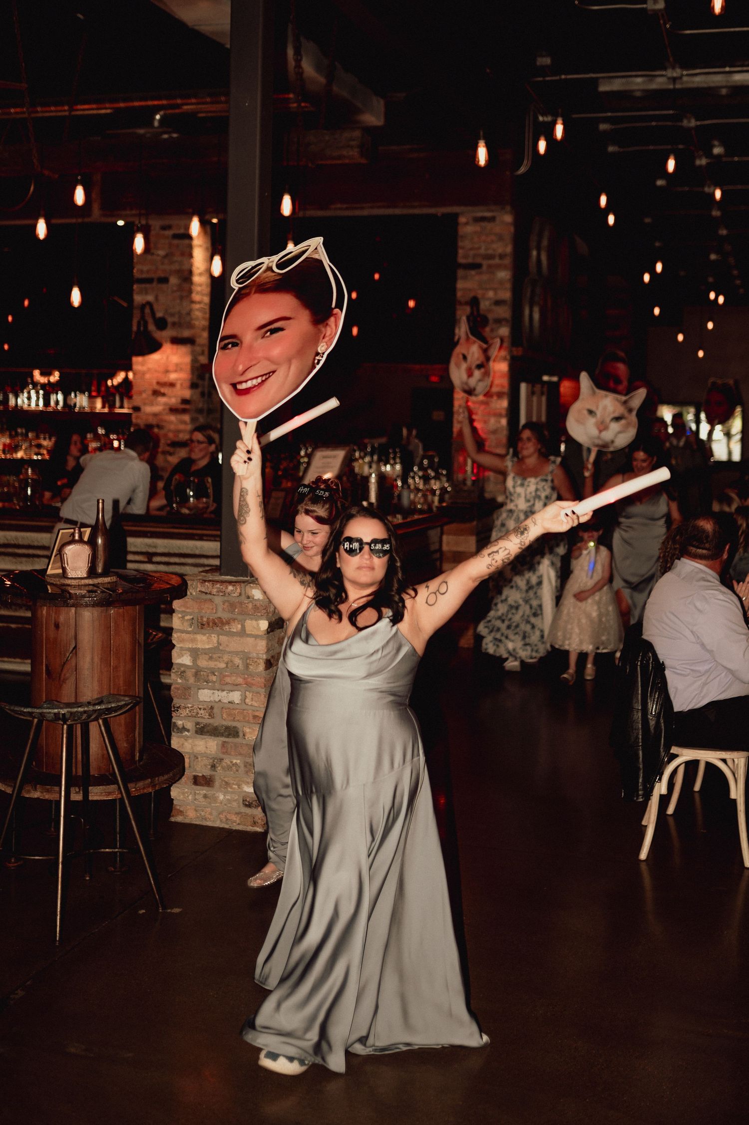 A person in a light gray dress dances joyfully in a dimly lit venue with string lights overhead.