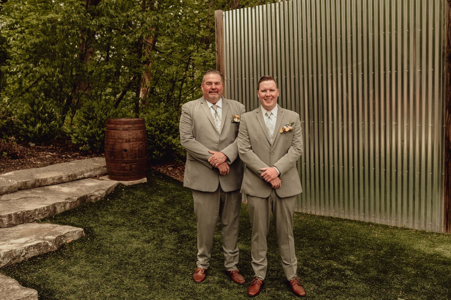Two people in light gray suits stand together in an outdoor garden setting with corrugated metal wall behind them.